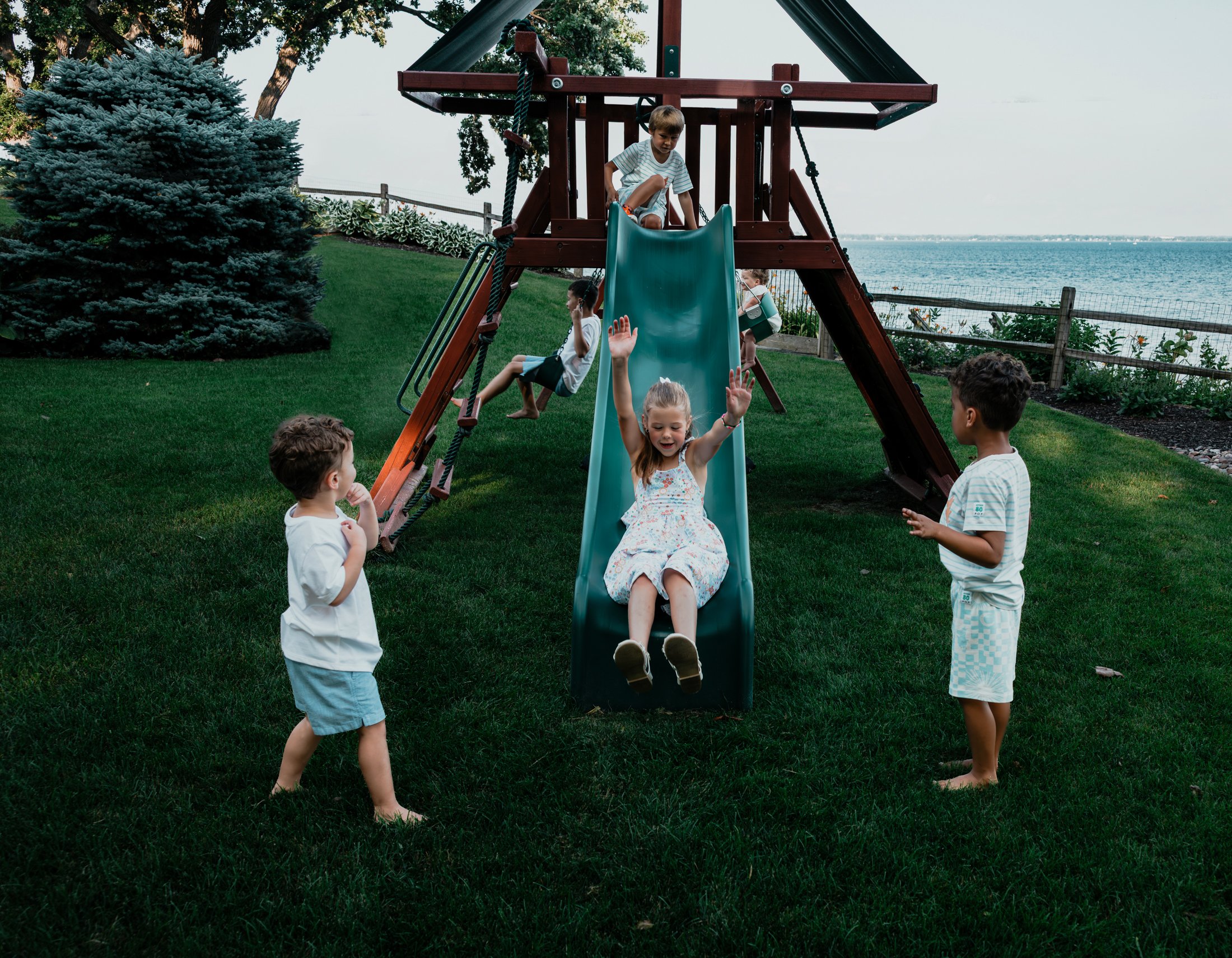 Children playing on a backyard playground near water, with a girl sliding down and other kids waiting and watching.