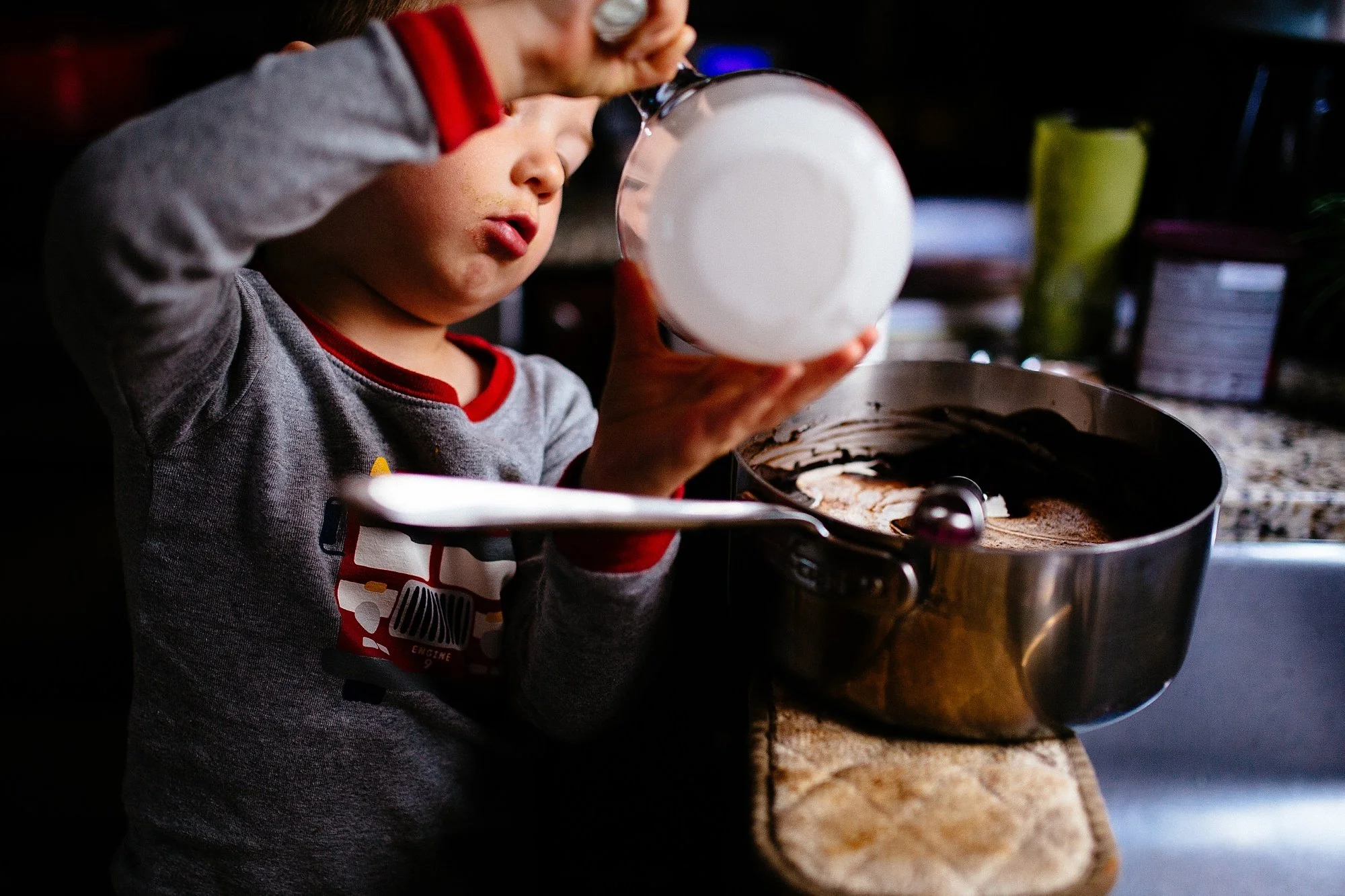A young boy wearing a gray long sleeve shirt with red trim is pouring milk into a bowl of chocolate cake batter. There is a kitchen countertop with various items in the background.