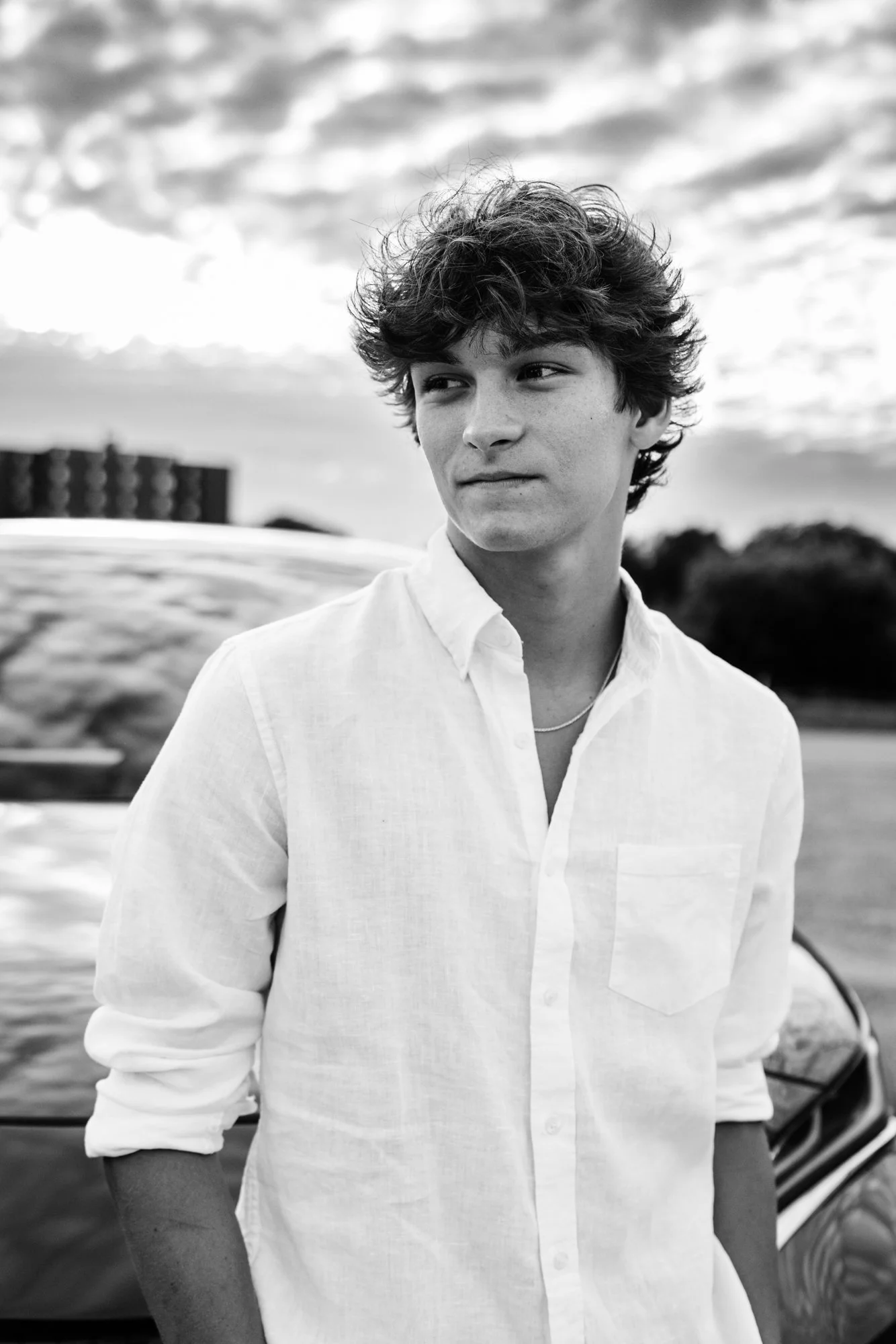 Black and white photo of a young man with curly hair, wearing a white shirt with sleeves rolled up, standing outdoors near a car, with a cloudy sky in the background.