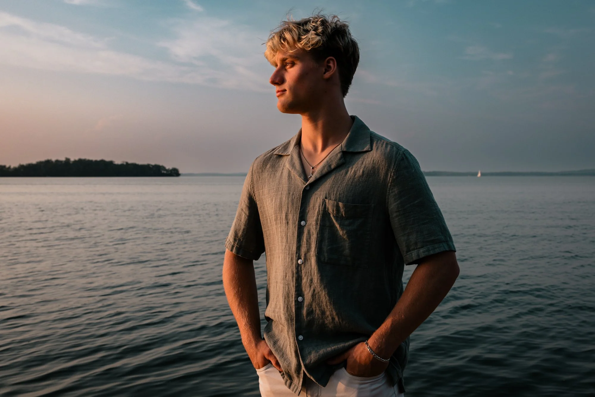 A young man standing by the water at sunset, looking to the left, with his hands in his pockets.