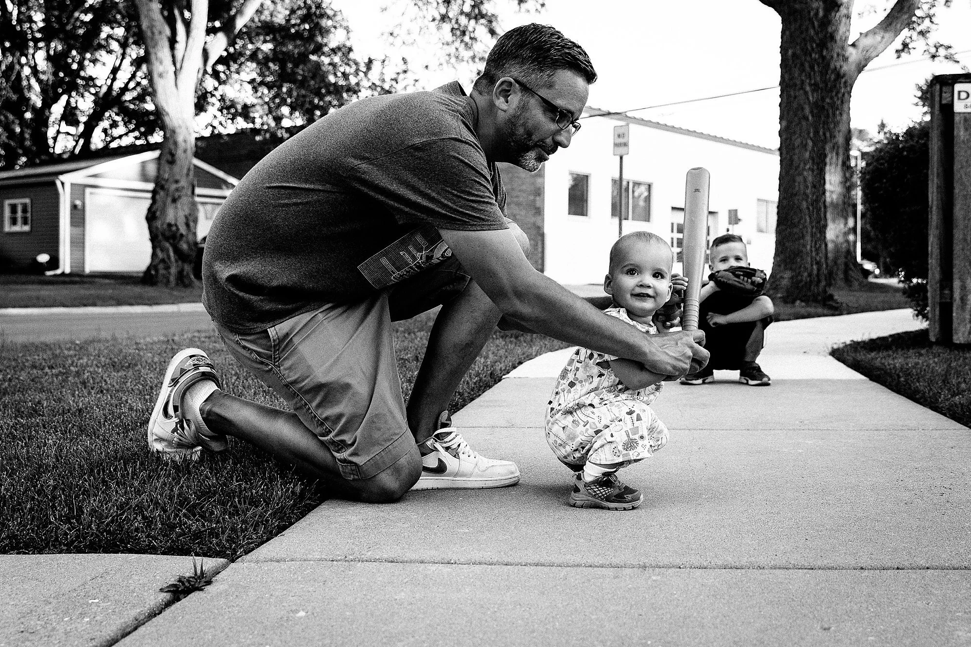 A man kneeling on the grass helping a toddler learn to walk with a baseball bat in hand. In the background, a young boy squatting with a baseball glove. Trees and houses in the background.