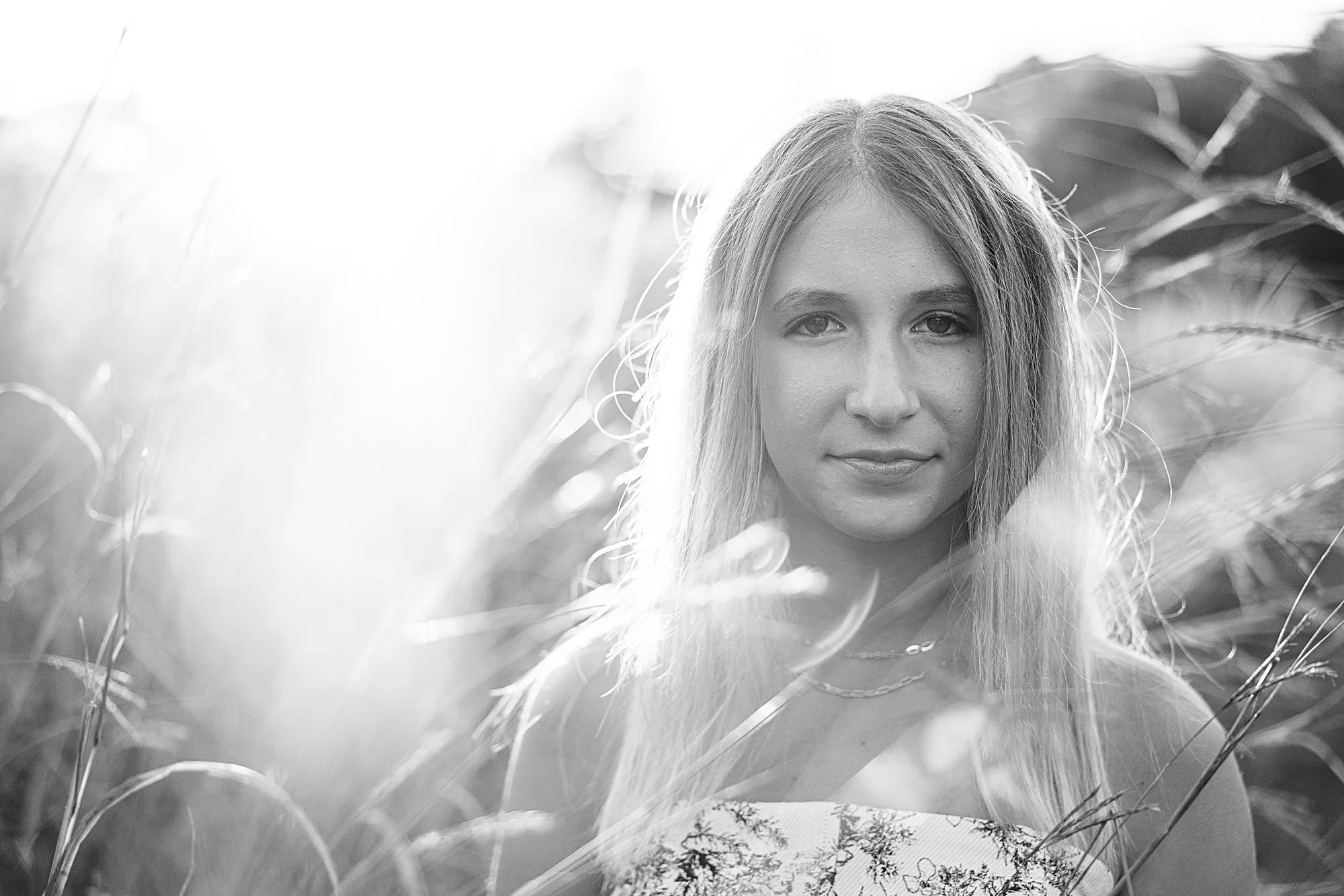 A black and white photo of a young woman with long hair, standing amidst tall grass, with sunlight shining from behind her.