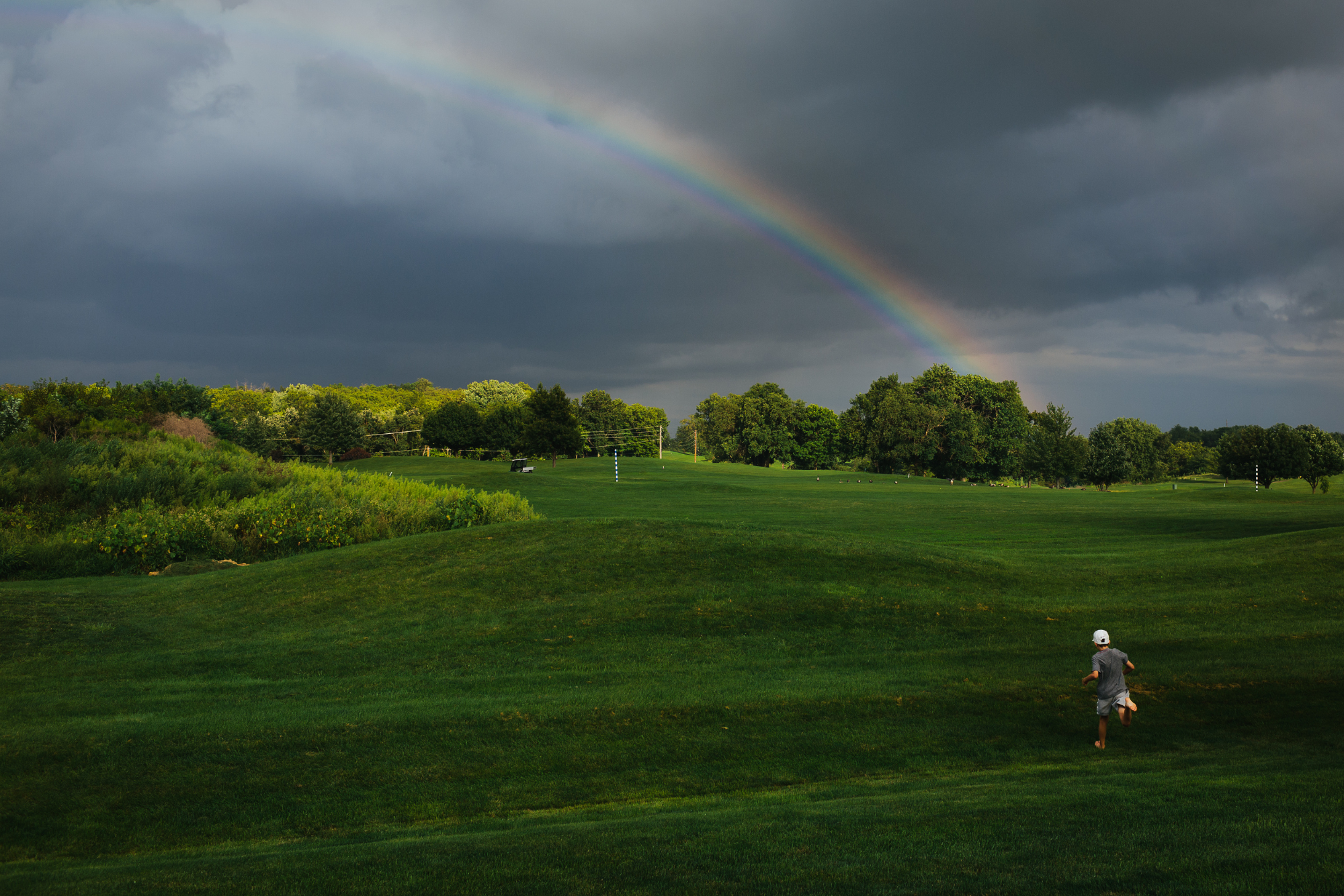 A person running on a lush green golf course during a storm, with dark clouds and a rainbow in the sky.