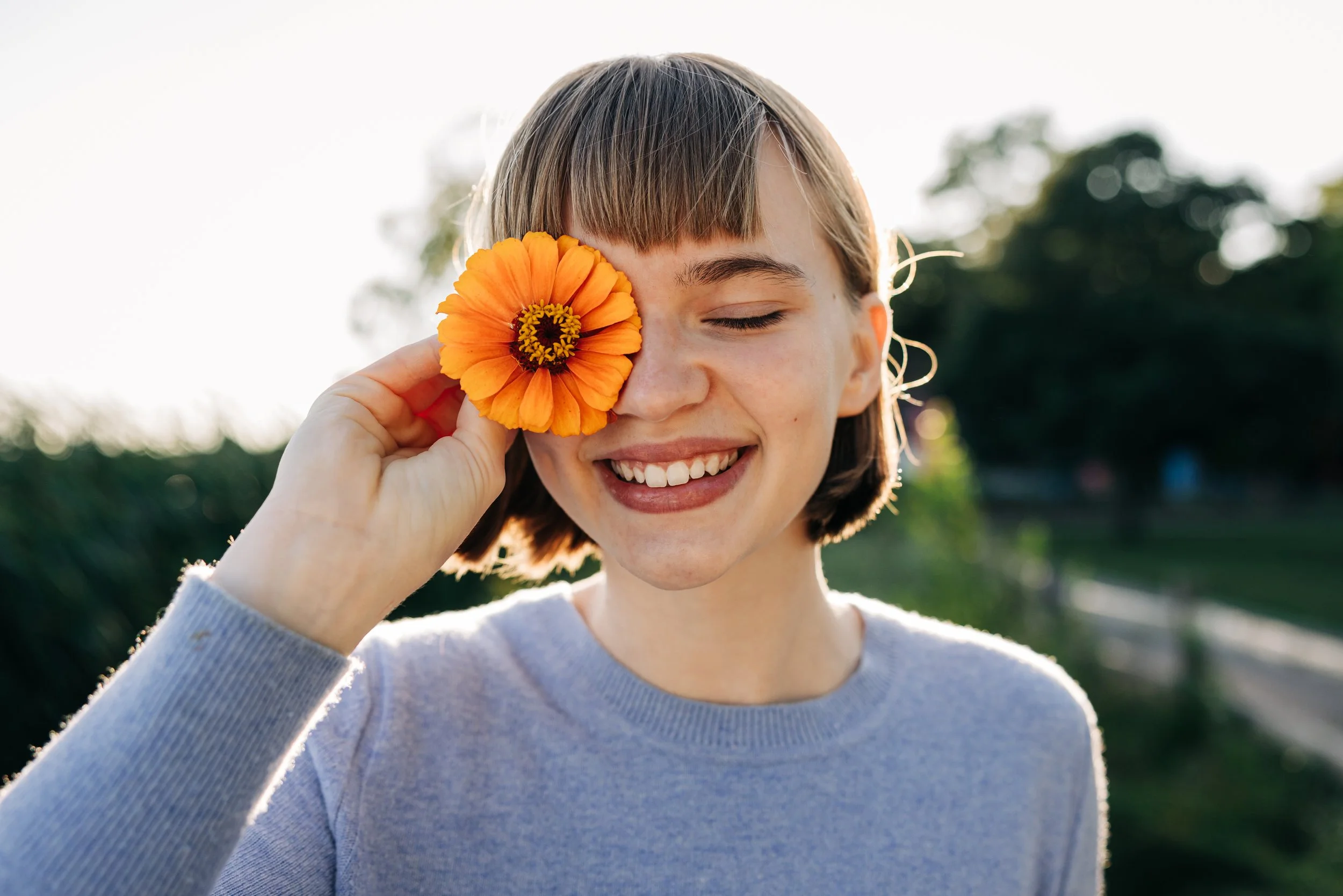 A woman with short brown hair smiling with eyes closed, holding an orange flower over her eye, outdoors with trees in the background.