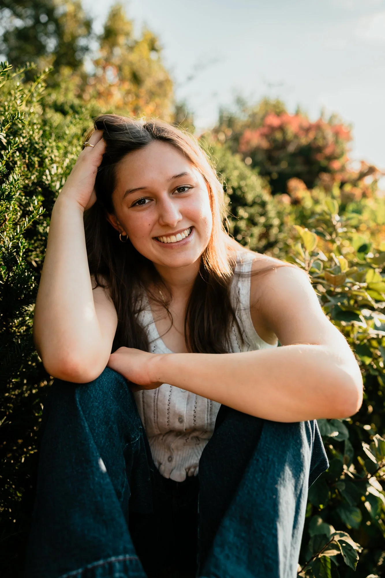 A young woman sitting outdoors among green bushes, smiling and looking at the camera, with her left hand resting on her knee and her right hand on her head, during golden hour sunlight.