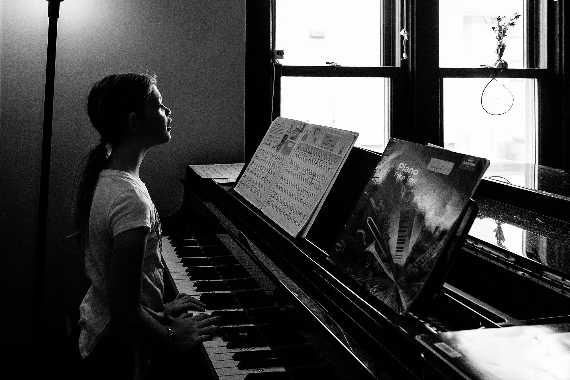 A young girl sitting at a piano, reading sheet music, with sunlight coming through a window behind her.