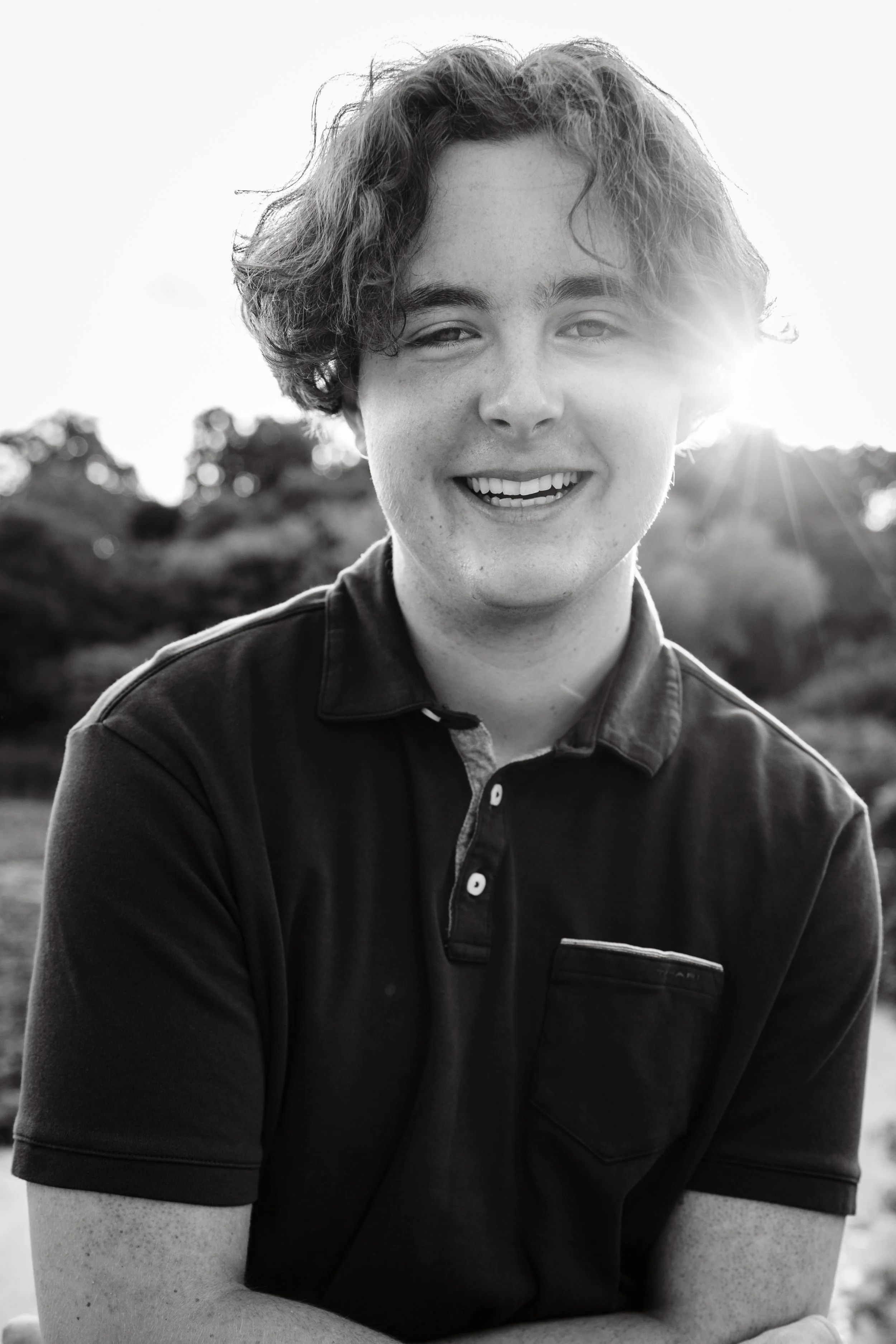 A smiling young man with curly hair in black and white, outdoors with sunlight in the background.