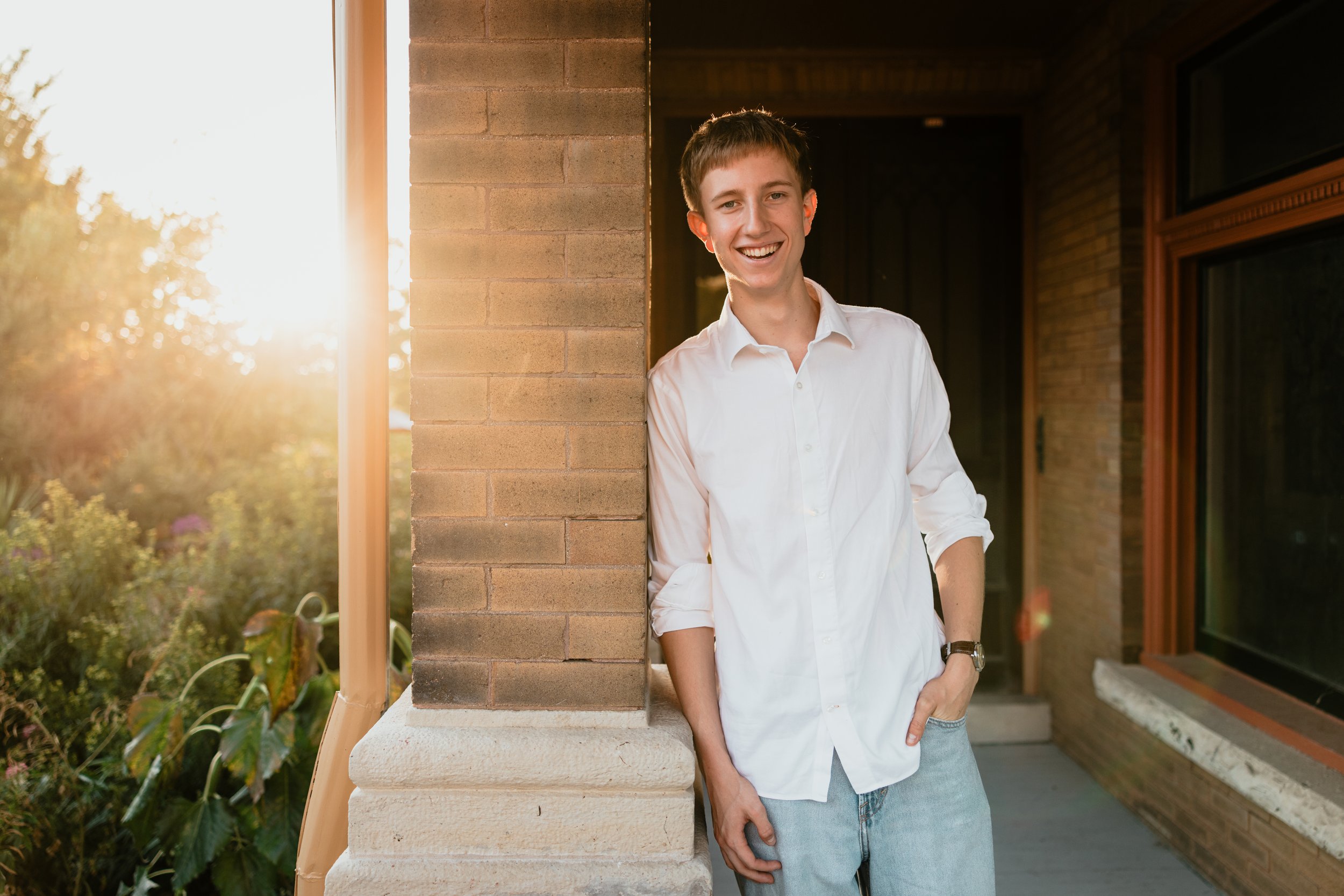 A young man in a white shirt and light jeans standing outdoors next to a brick column, smiling at sunset with sunlight streaming in the background.