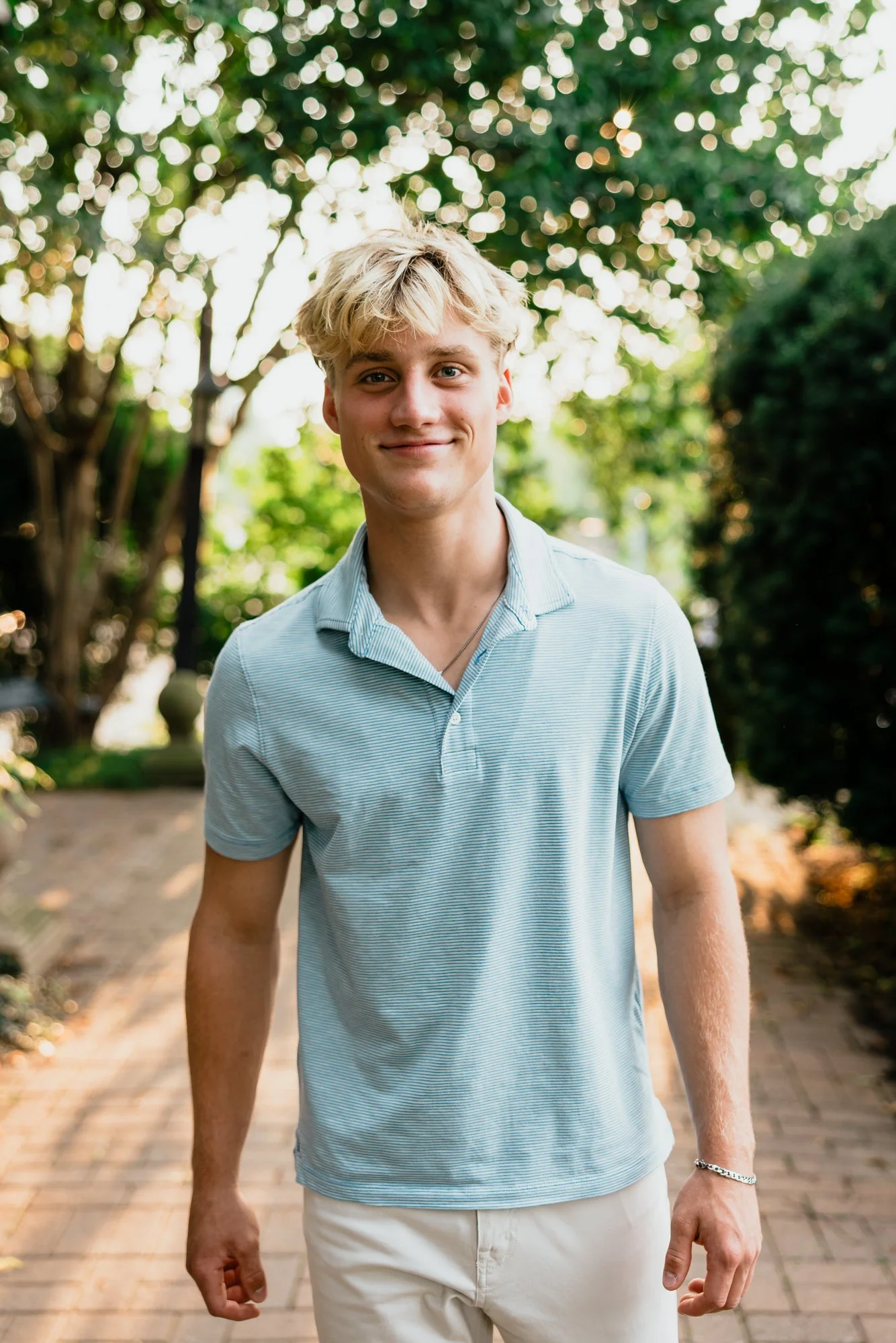 A young man with blonde hair wearing a light blue polo shirt and white pants standing outdoors on a sunny day with greenery in the background.
