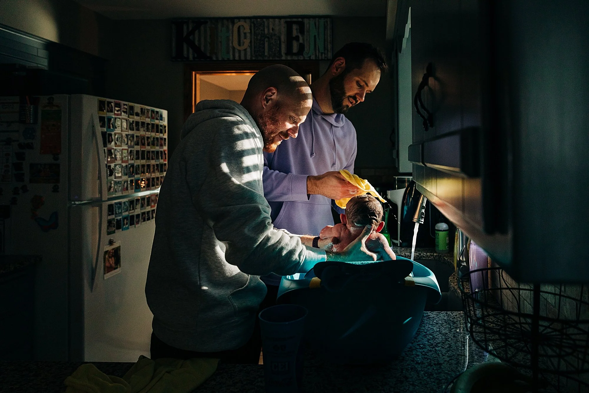 Two men are giving a bath to a baby in a kitchen. The man on the left is wearing a gray hoodie, and the man on the right is wearing a purple hoodie. Sunlight is casting shadows through blinds onto the scene.