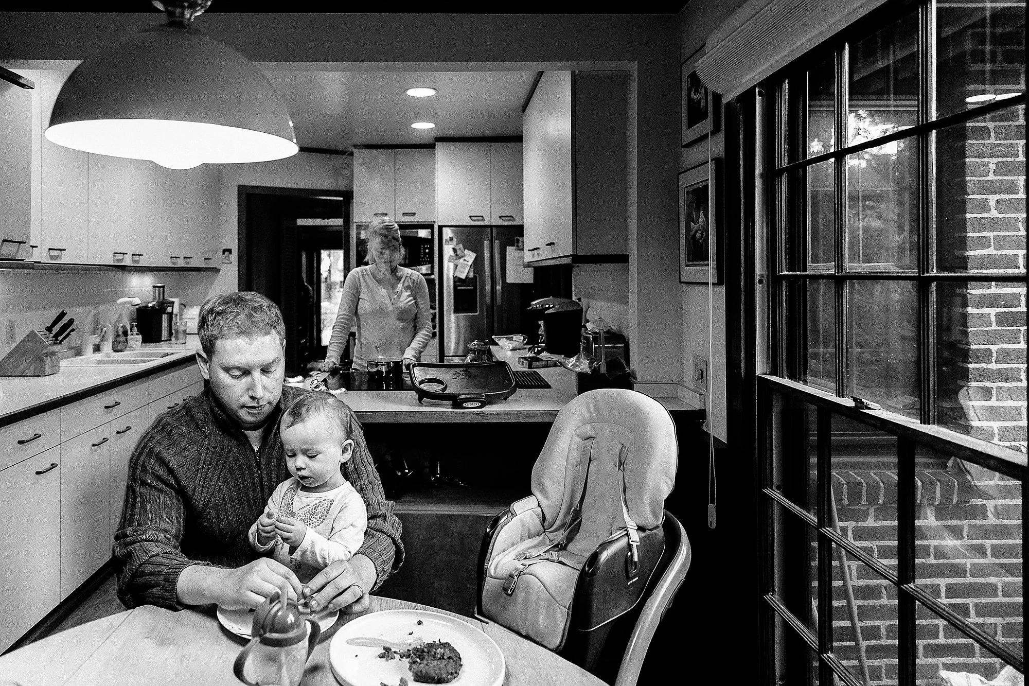 A family in a kitchen with a man and a young girl at the dining table, a woman preparing food in the background, and a large window showing brick exterior walls.