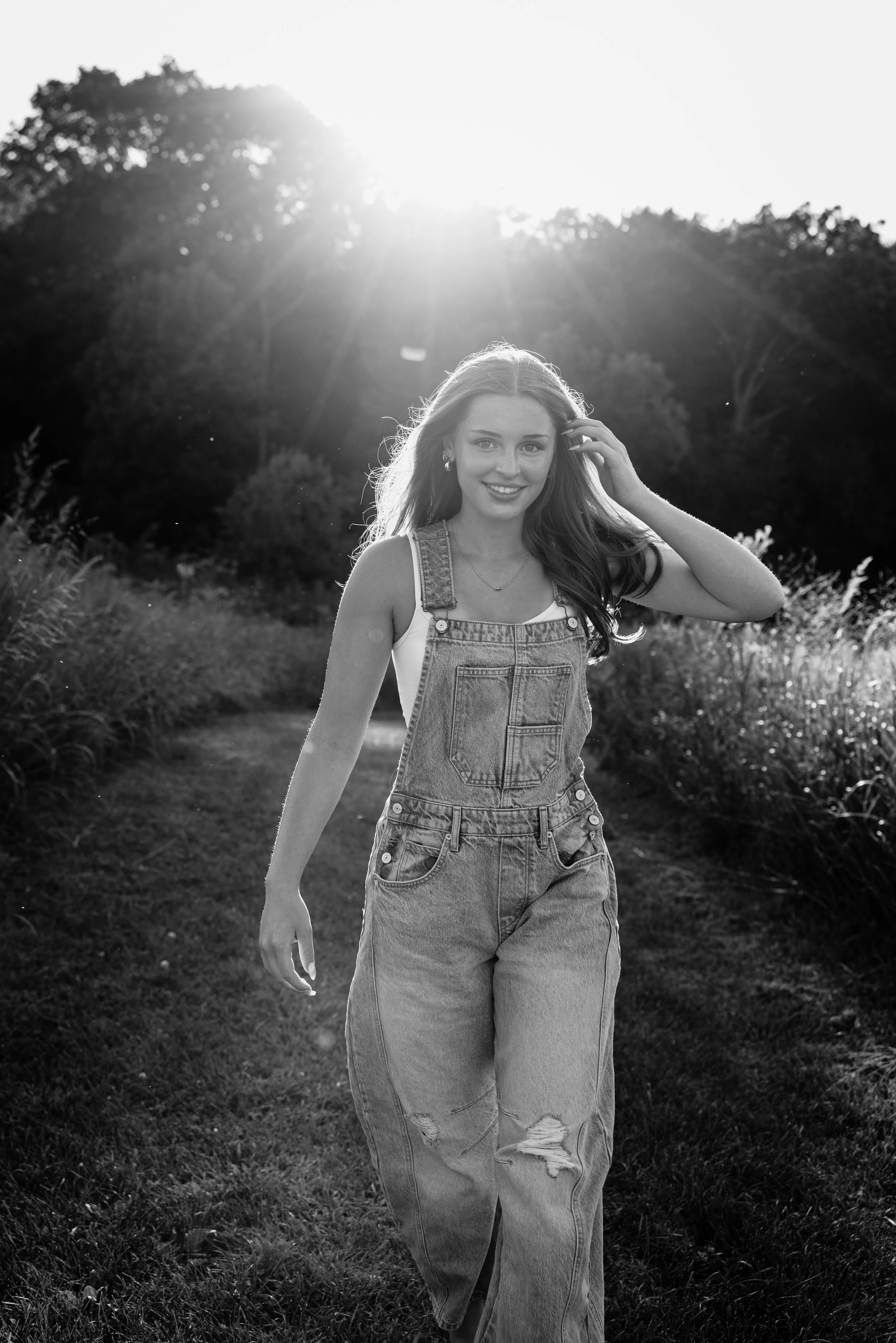 A young woman walking outdoors on a grassy path, smiling, with sunlight shining behind her, in a natural setting with trees and bushes.