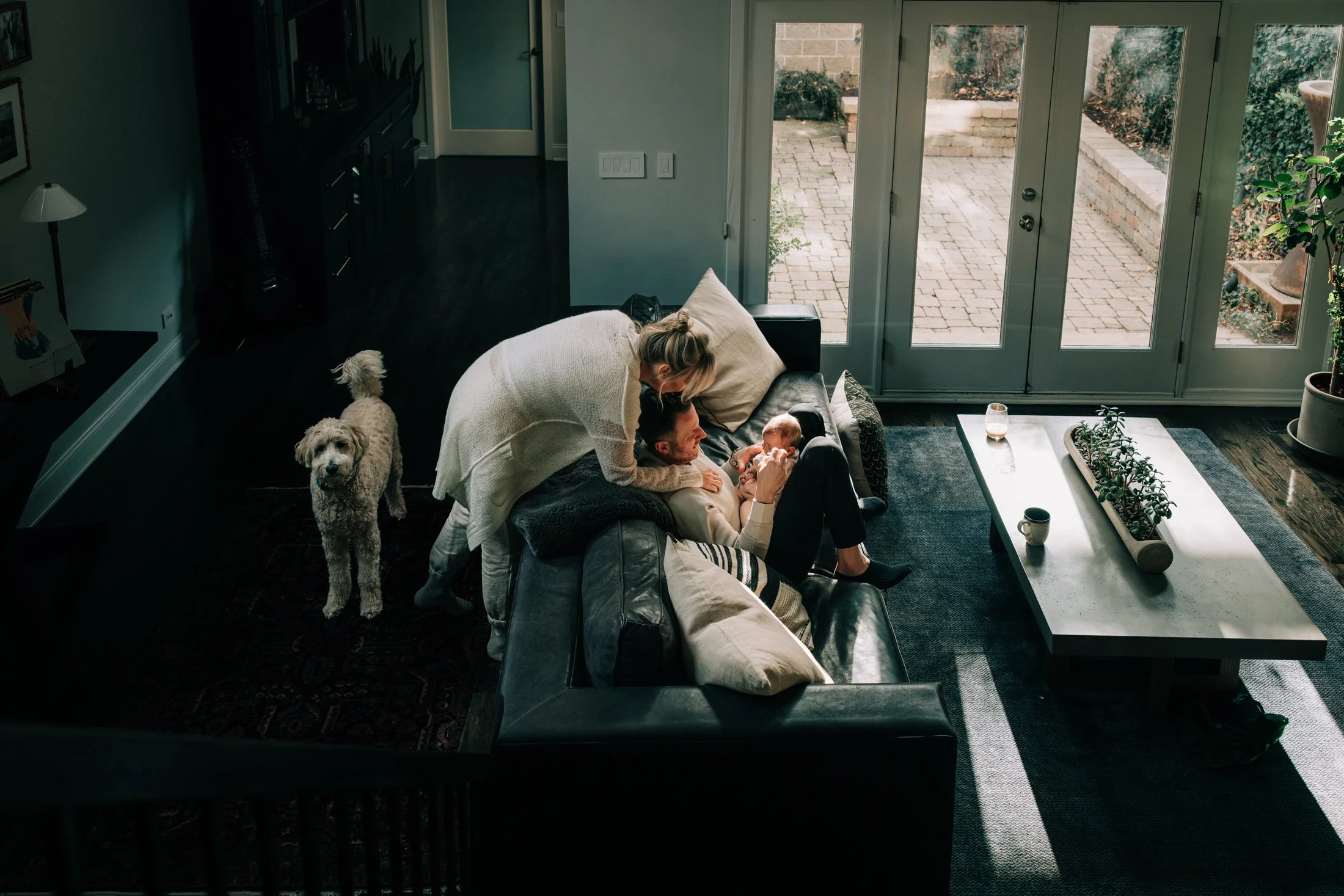 A woman and a man sitting on a black leather couch, with the woman leaning over the man and holding a small baby, inside a living room with a French door leading outside. A white dog is standing nearby on a dark rug, and a coffee table with plants, a
