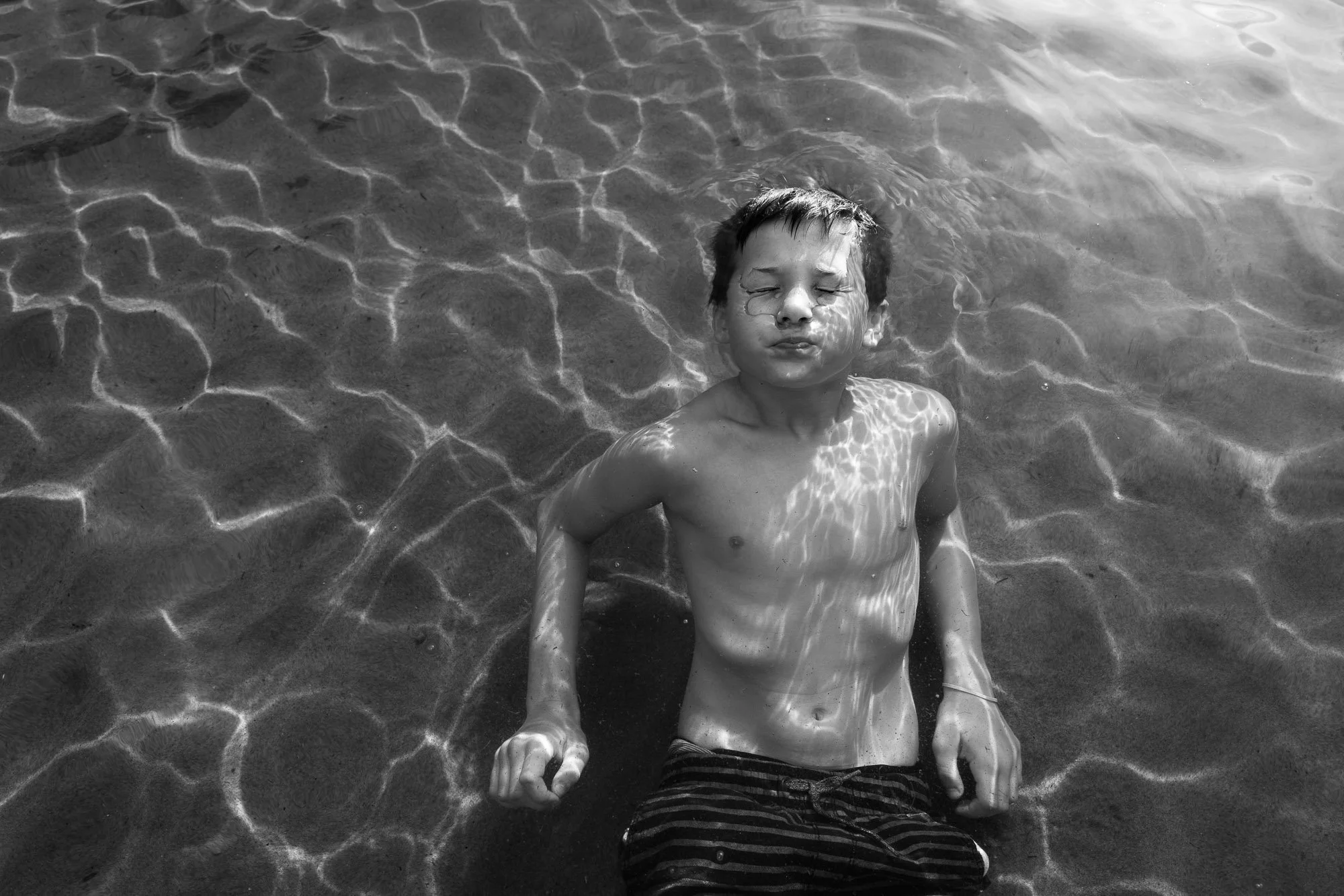 A young boy with closed eyes appears to be relaxing in shallow water at the beach, with light reflecting off the water's surface.