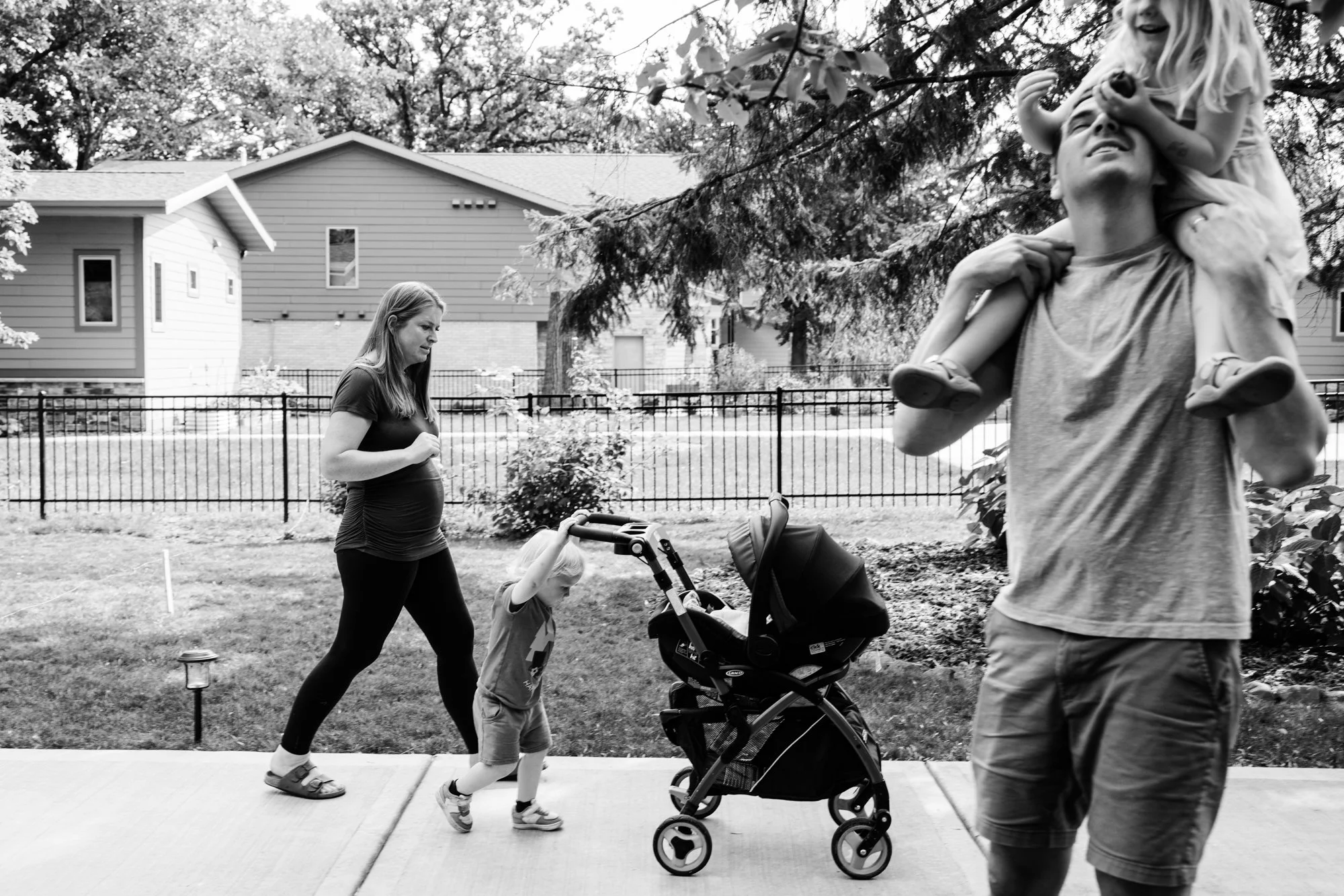 A family walking outdoors near a suburban house. A pregnant woman is pushing a stroller, a young girl is sitting on an adult man's shoulders, and an adult man is walking alongside them on a sidewalk.