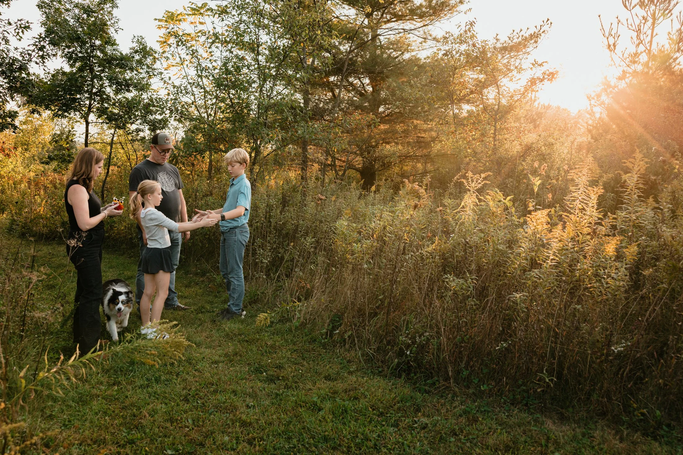 A family is standing outdoors on a grassy path during sunset, with tall grass and trees in the background. The family includes two adults, two children, and a dog. The children are exchanging something, and the adults are watching or holding items.