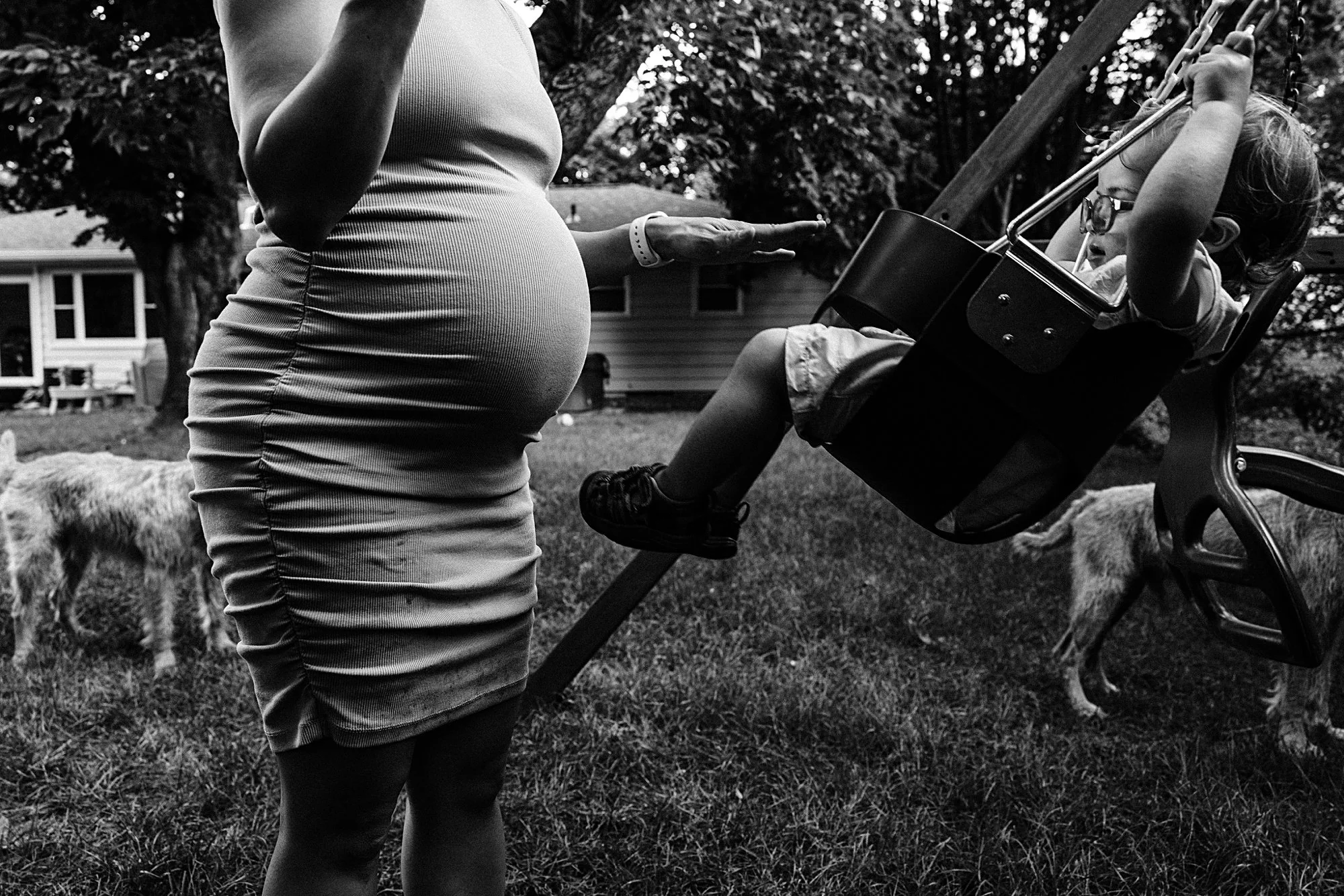 A pregnant woman stands outside in a yard, reaching out her hand towards a young girl in a swing seat. The girl wears glasses and has her arms raised, holding onto the swing's chains. There are trees, a house, and some dogs in the background.