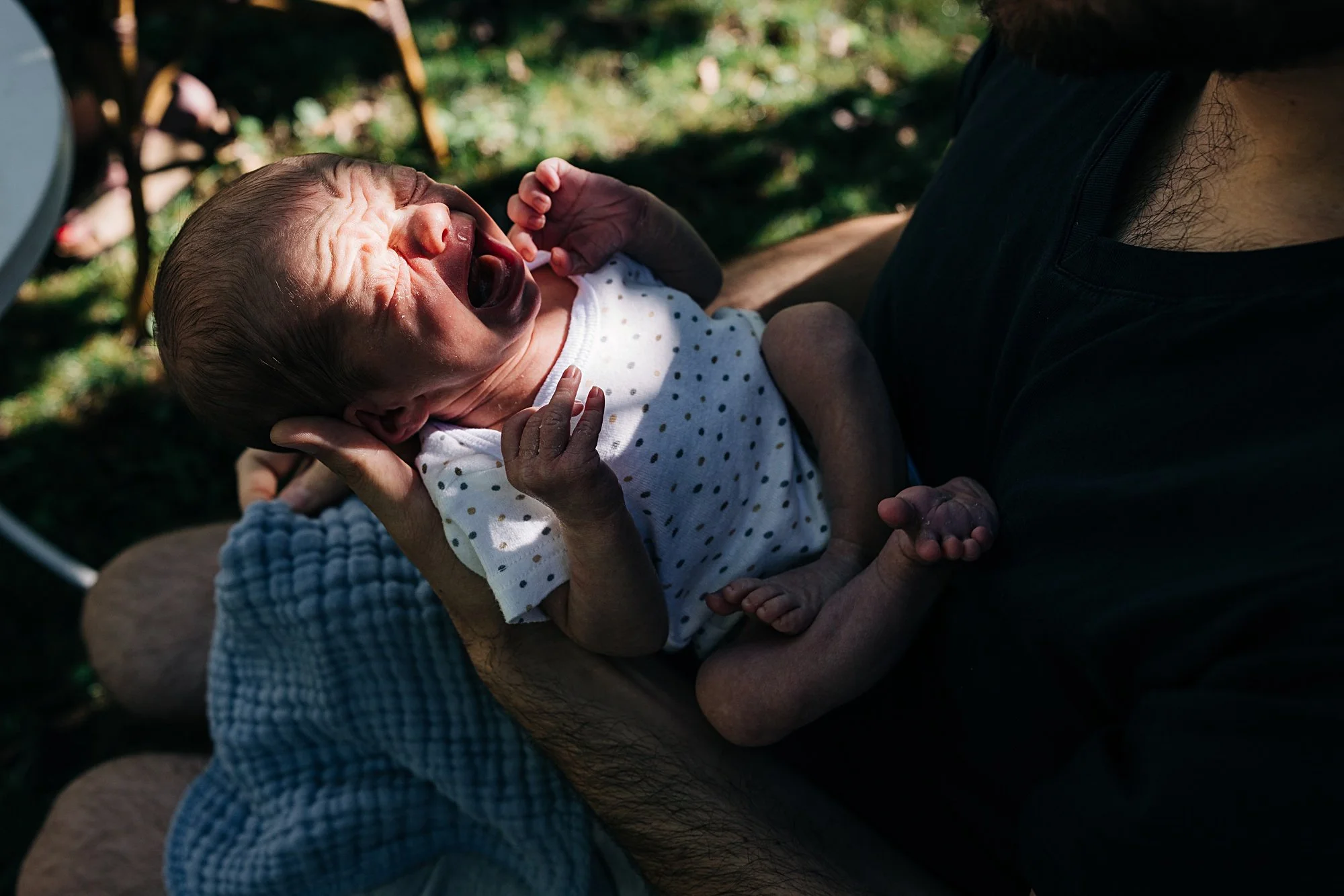 A newborn baby crying while being held by a person outdoors in sunlight.