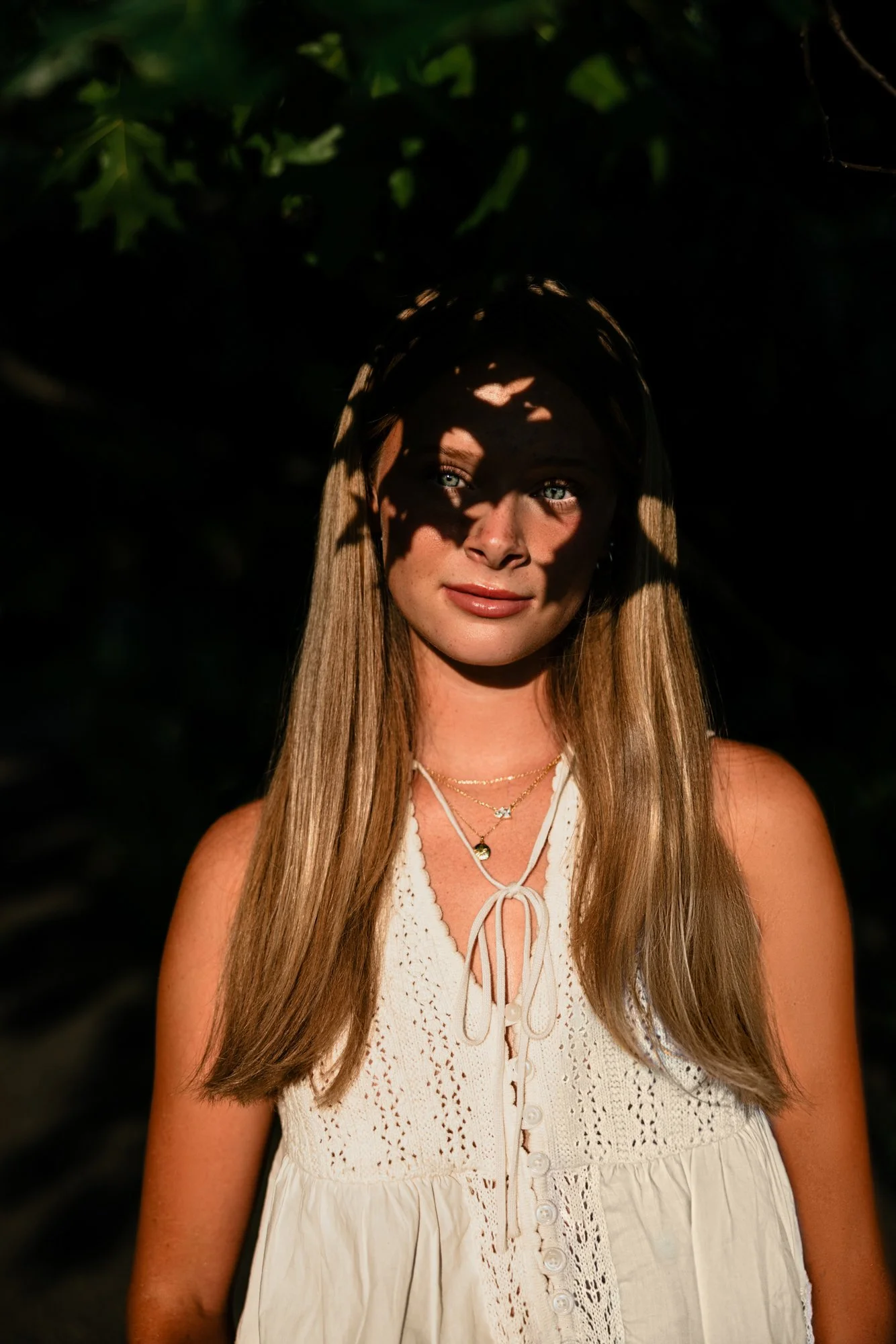 A woman with long, light brown hair and blue eyes, wearing a white sleeveless top with lace details, standing in a dark outdoor setting with patterns of shadow and light on her face caused by leaves.