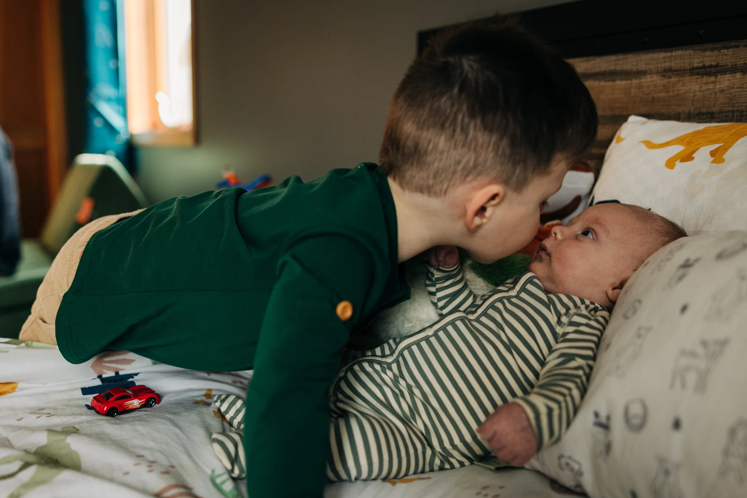 A young boy leans over to kiss a newborn baby lying on a bed.