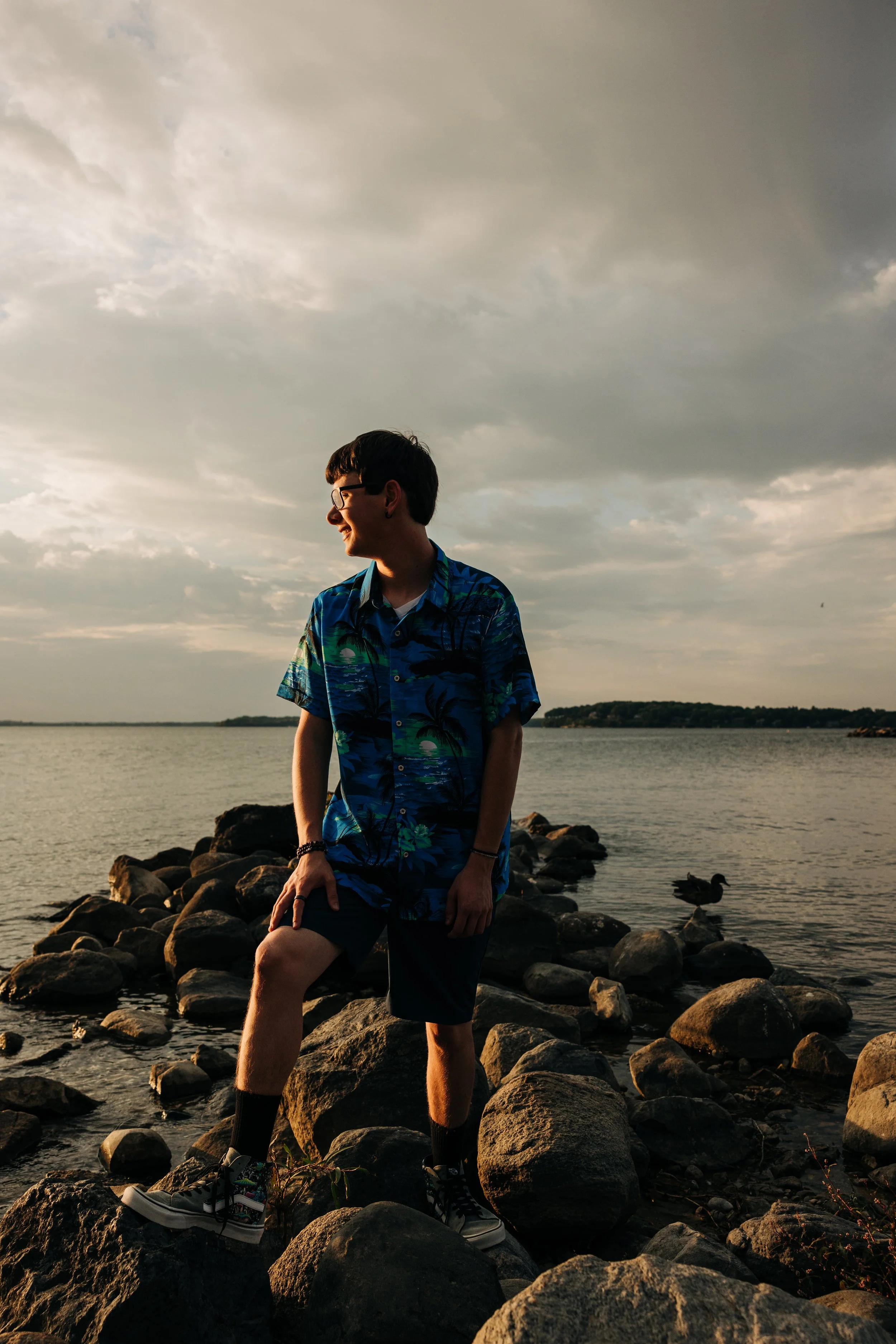 A young man wearing a colorful blue Hawaiian shirt, shorts, and sneakers standing on rocks by a lake at sunset, with clouds in the sky and a duck on the water.