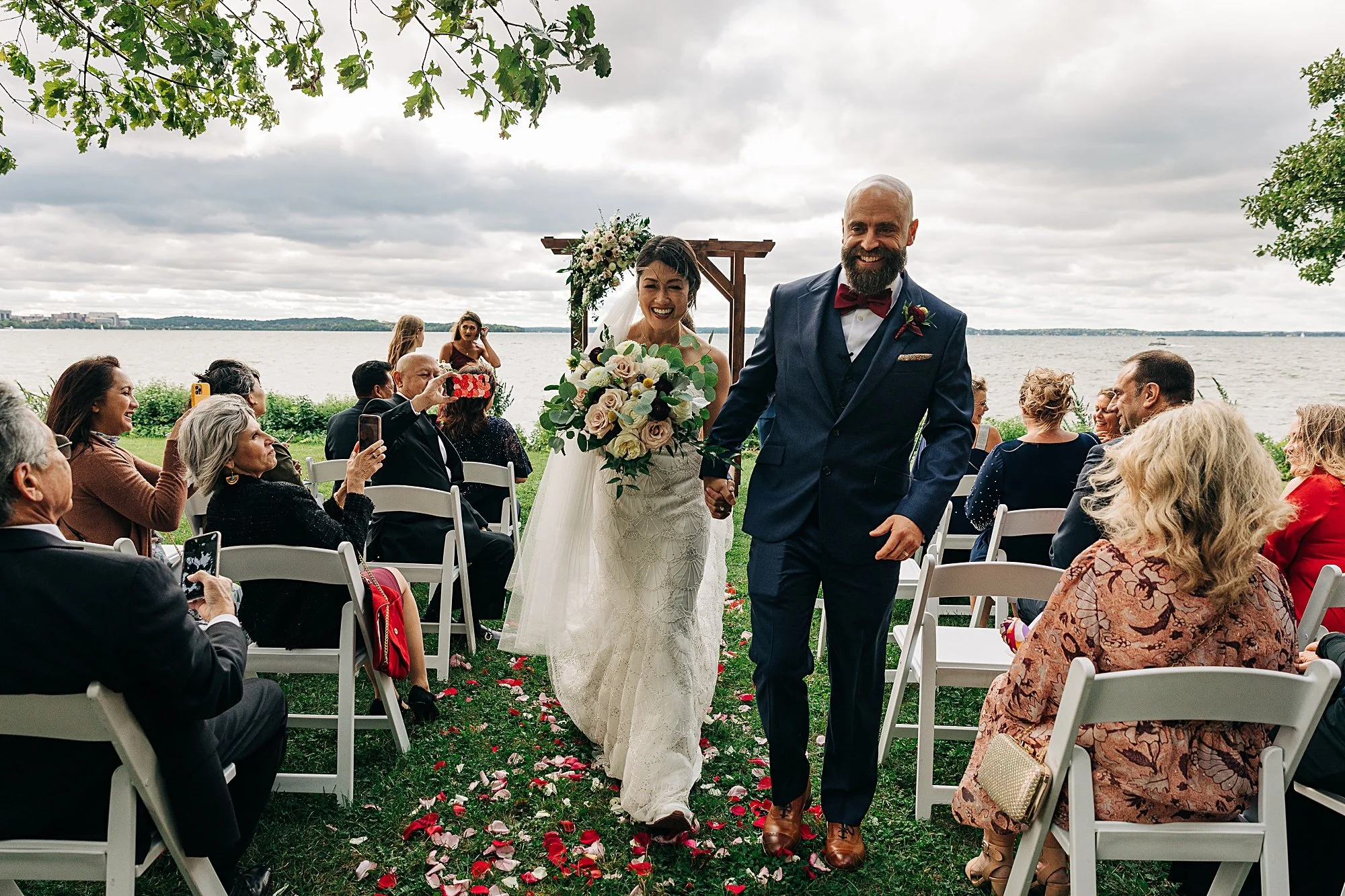 Bride and groom walking down the aisle at outdoor wedding near a lake, guests seated on white chairs, some taking photos, with a decorated wooden arch and cloudy sky in the background.