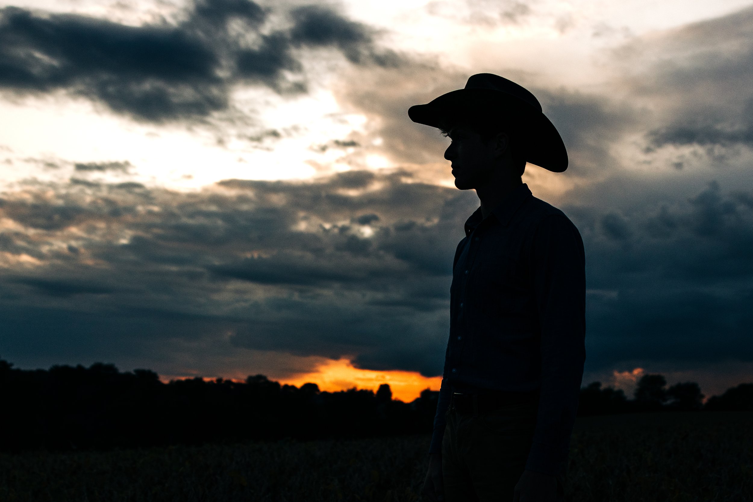 Silhouette of a man wearing a cowboy hat standing outdoors during sunset or sunrise with a cloudy sky in the background.