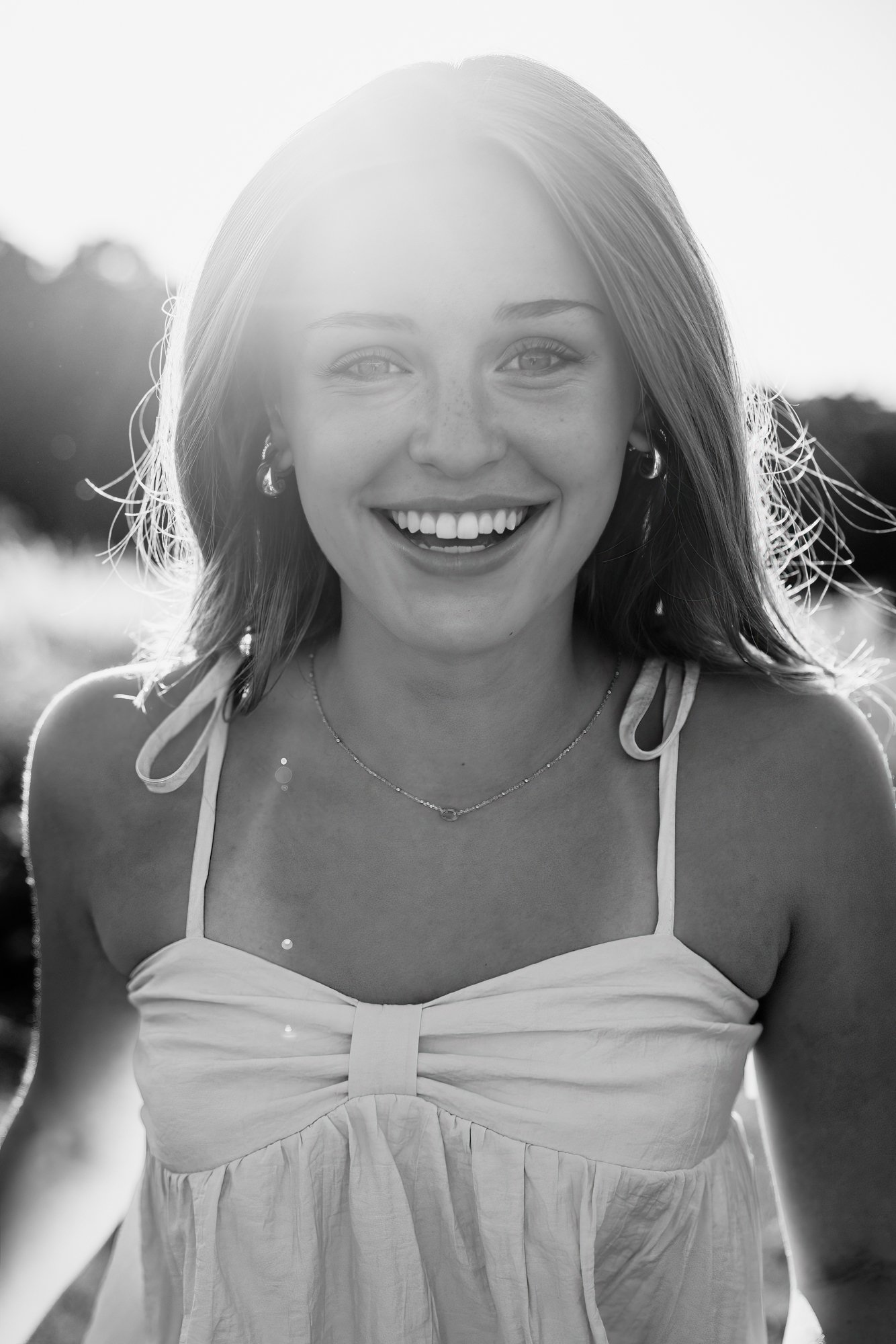 A smiling young woman with light skin, freckles, and shoulder-length hair, wearing a sleeveless dress and earrings, outdoors during daylight.