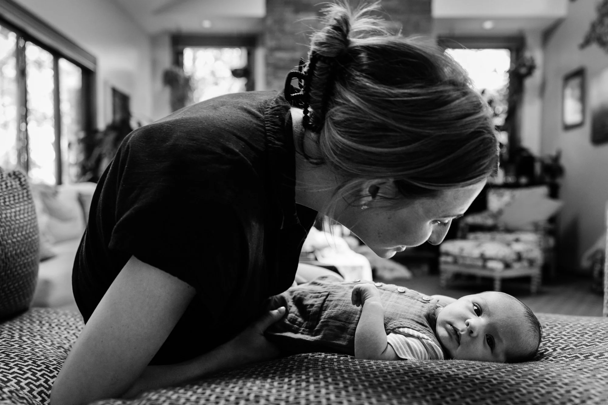A woman leaning over a baby lying on a patterned surface in a cozy living room, with natural light coming through the windows.