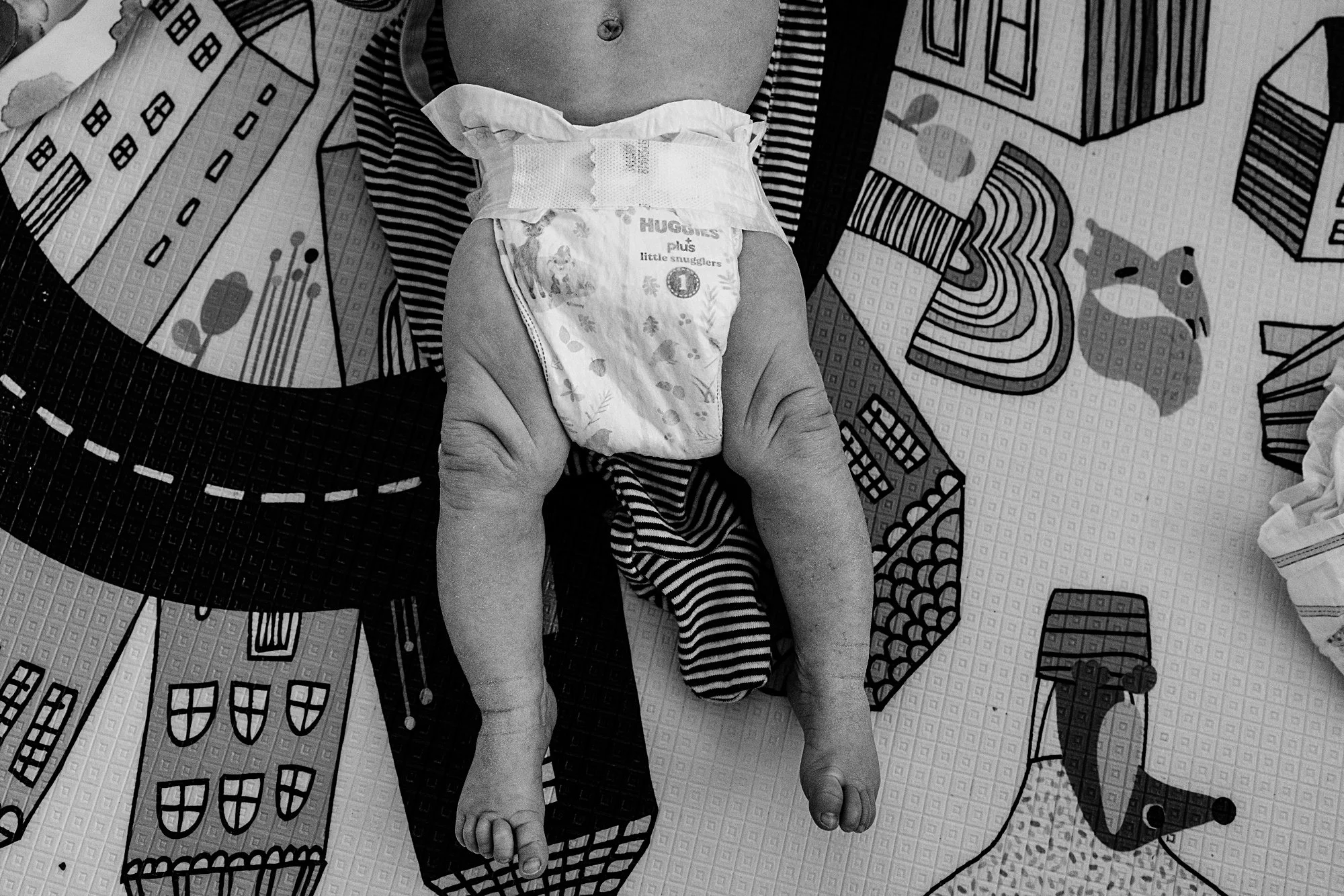 A baby lying on a colorful play mat, wearing a diaper and for the most part unclothed.