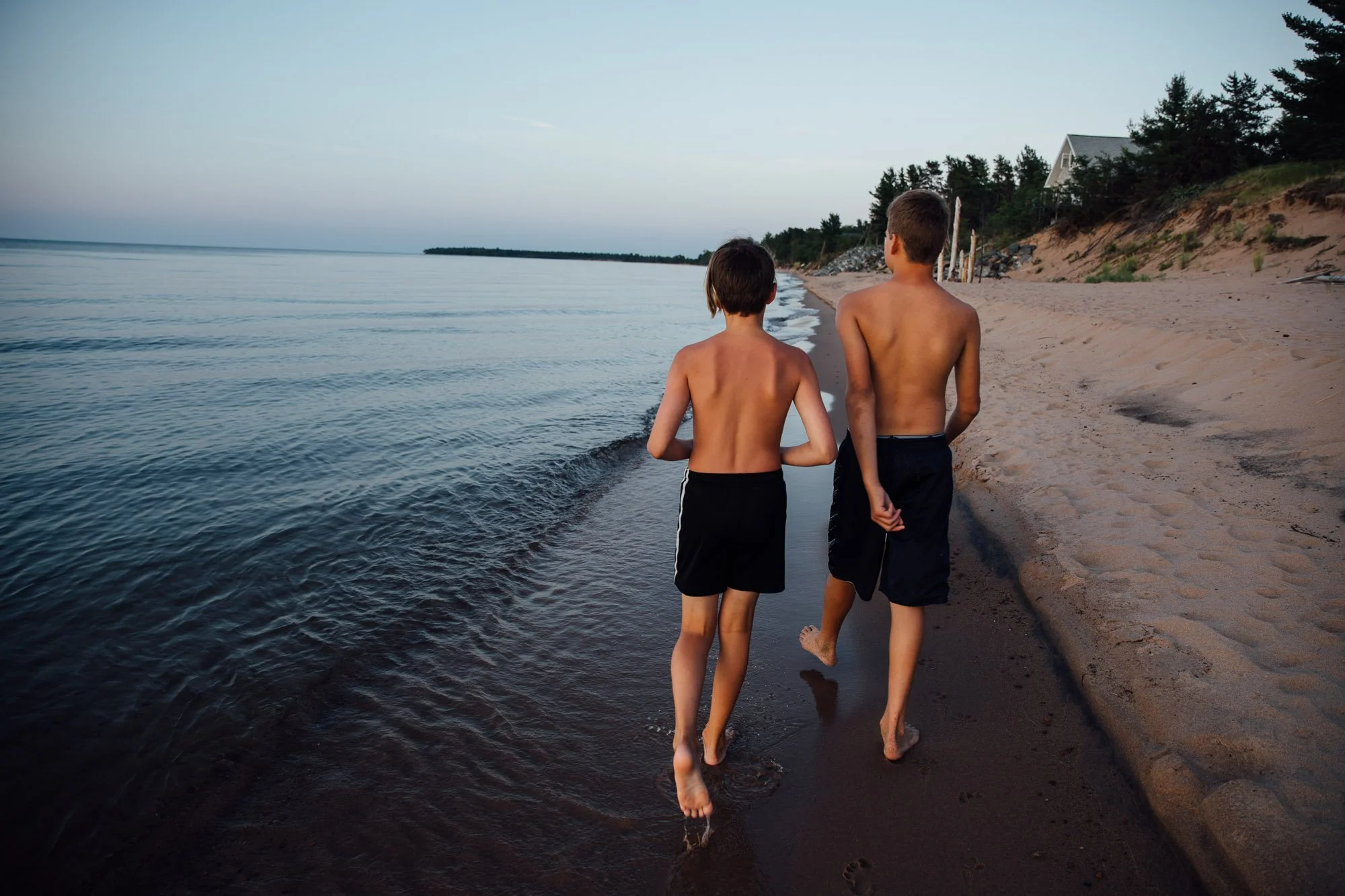 Two boys walk along the shoreline of a beach at sunset or early evening, with their backs facing the camera.