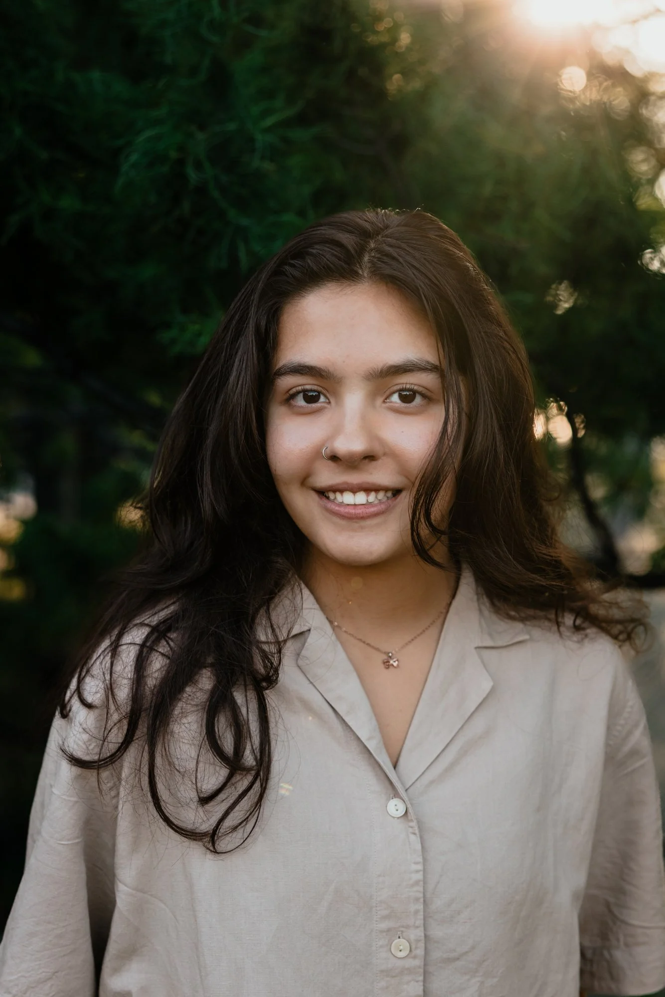 A young woman with dark hair and a nose ring smiling outdoors with trees and sunlight in the background.