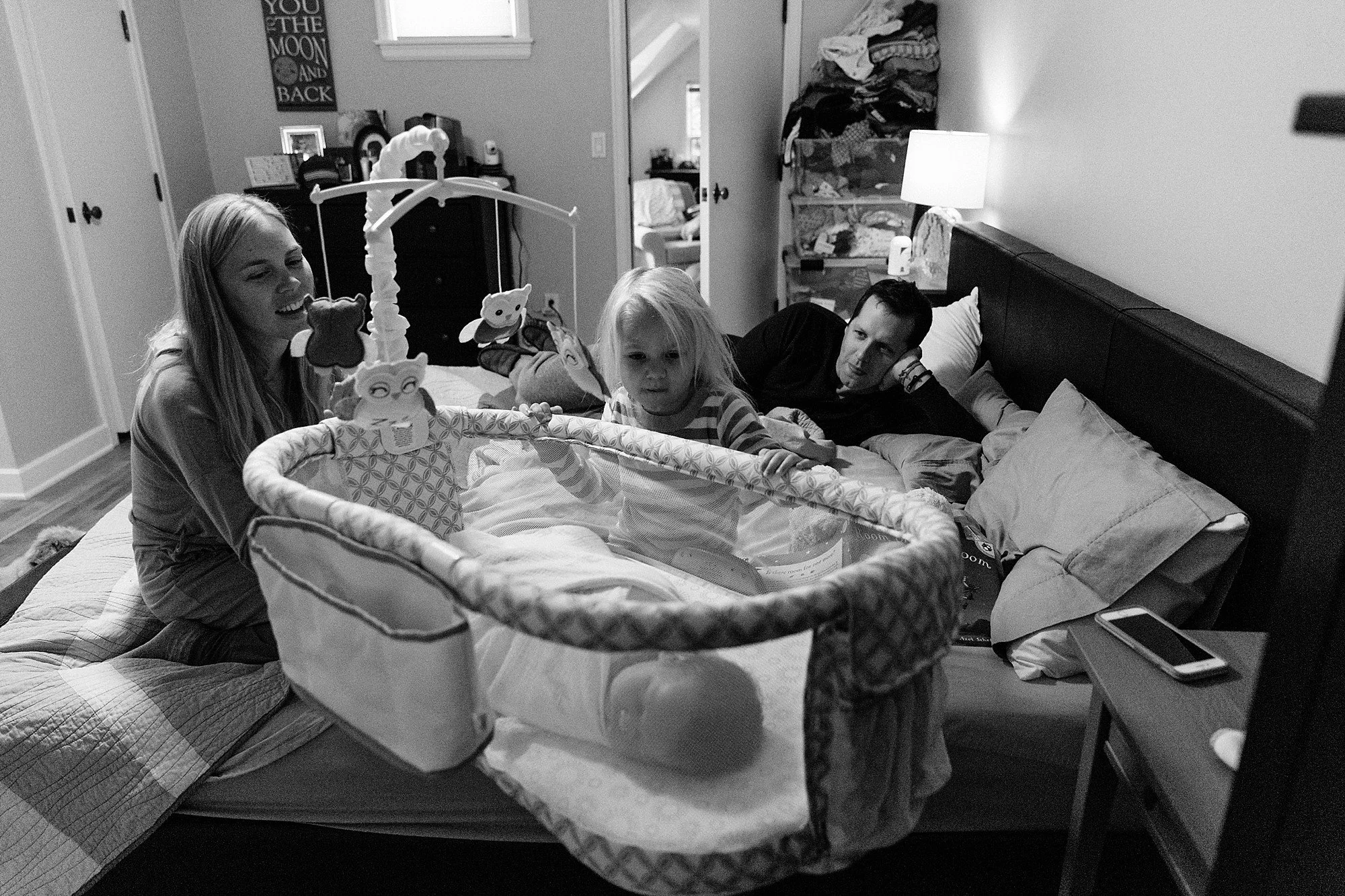 Mother, father, preschooler on a bed while overlooking large bassinet with tiny newborn inside