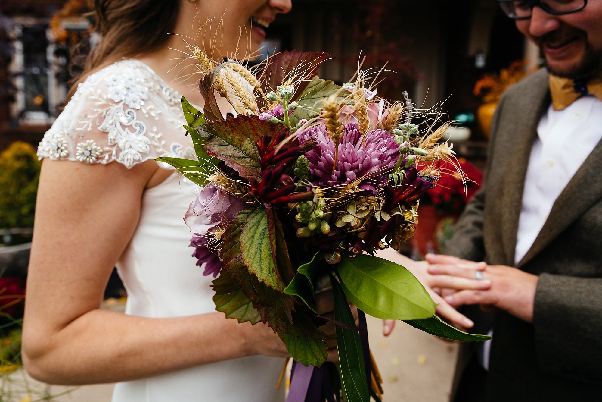 A bride holding a bouquet of purple, pink, and green flowers with leaves, while smiling at her partner during a wedding ceremony.