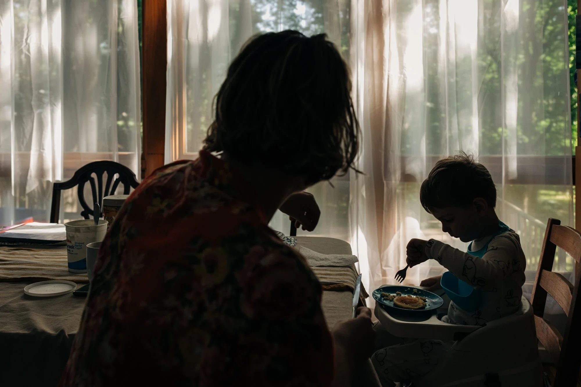 A woman and a young boy having a meal together at a dining table inside a room with large windows and sheer curtains, with a view of trees outside through the windows.