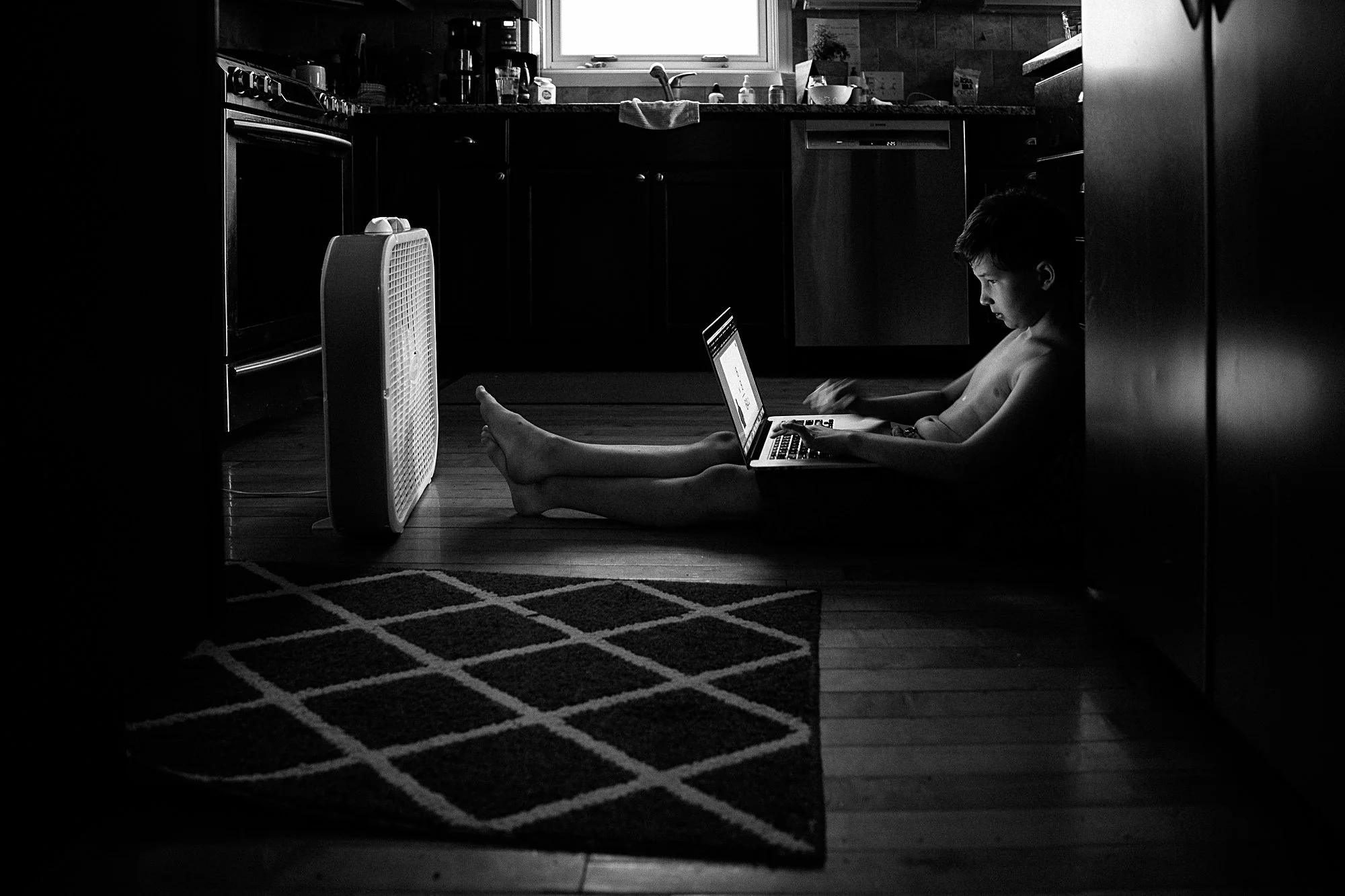 A young boy sitting on the kitchen floor using a laptop with a fan nearby, in a dimly lit kitchen with dark cabinets and a small rug in front.