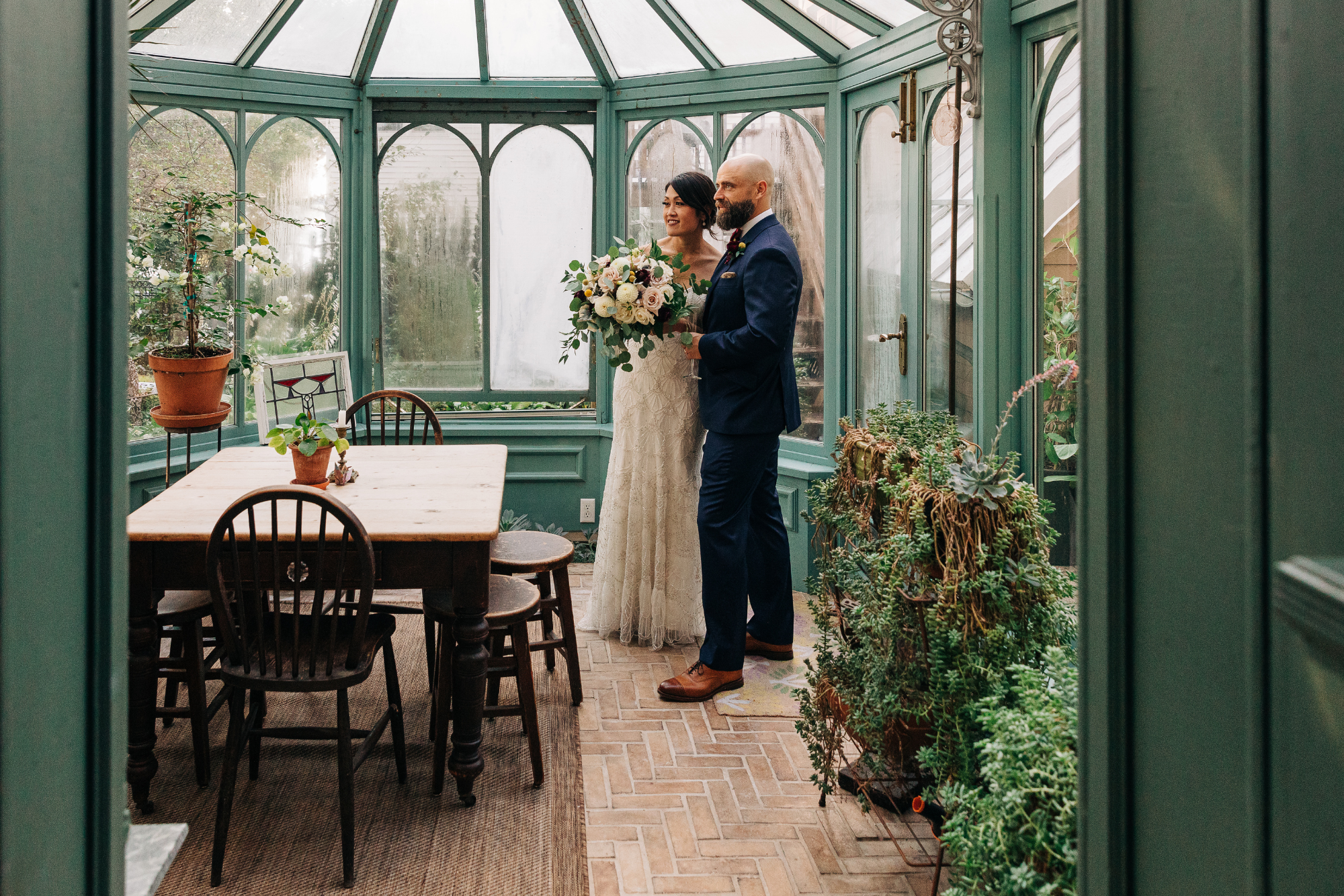 A bride and groom standing inside a green glass conservatory, the bride holding a bouquet of flowers and the groom in a navy suit, surrounded by indoor plants and wooden furniture.