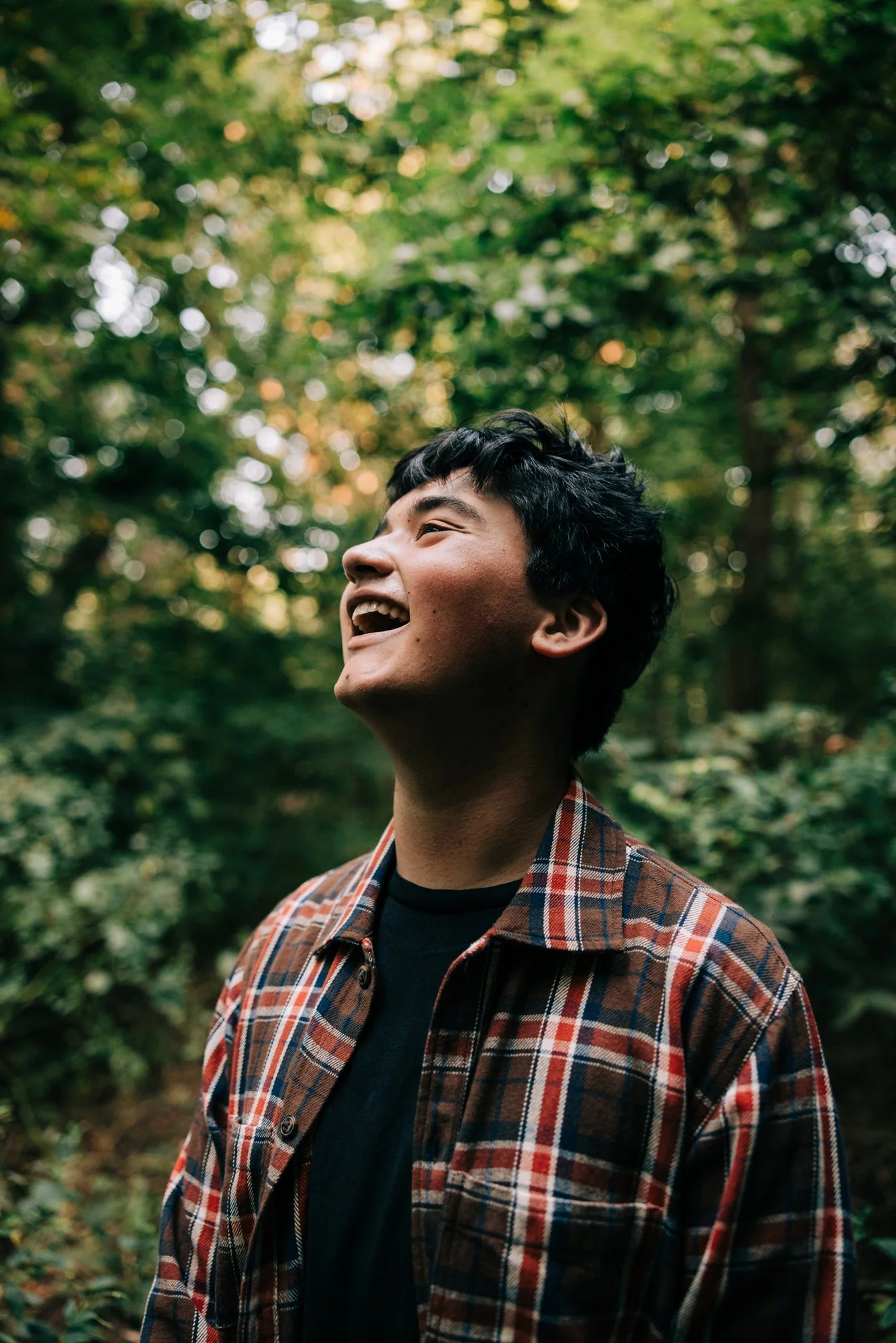 Smiling young man with black hair in a plaid jacket outdoors in a green, leafy forest.