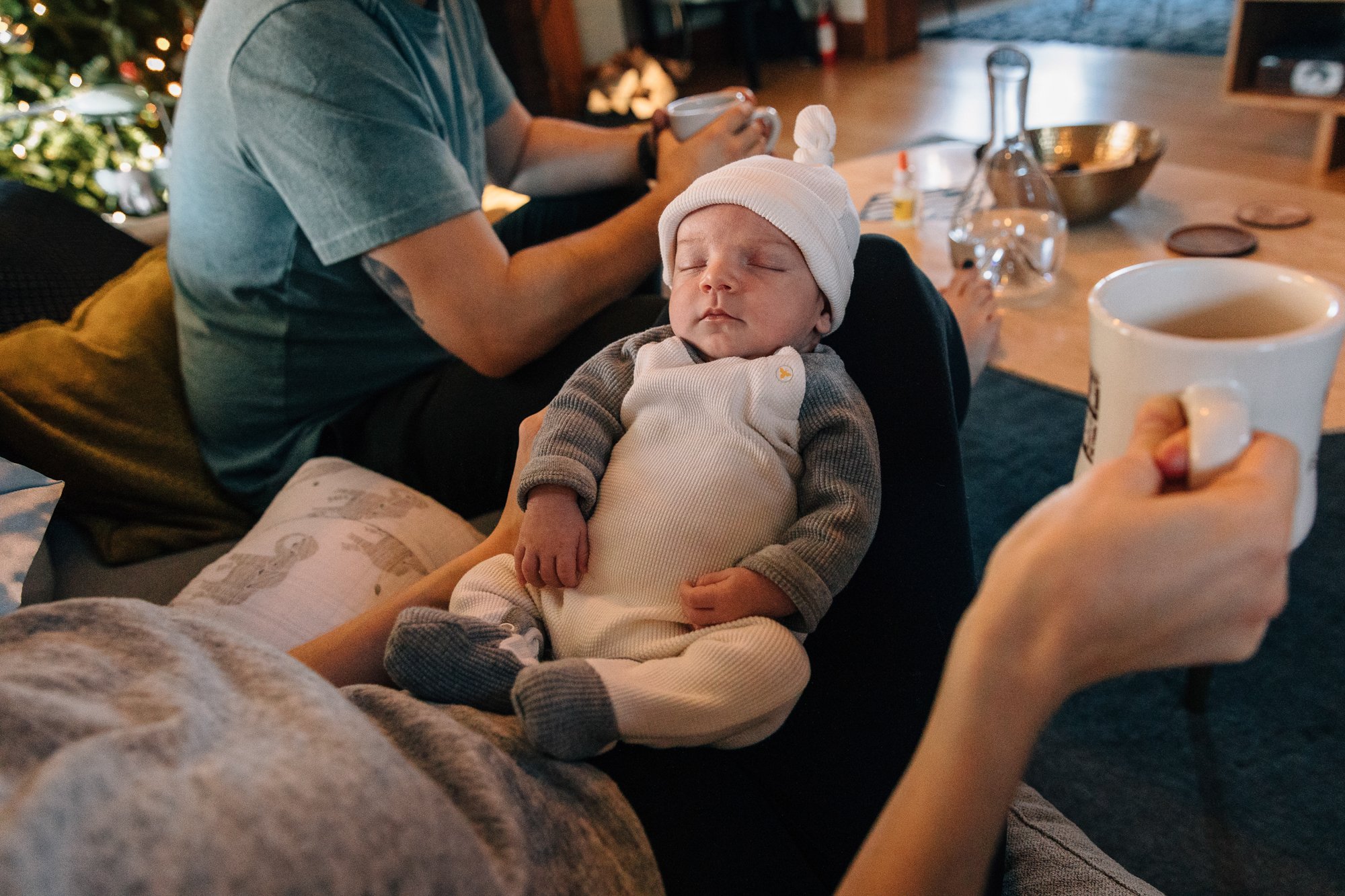 A sleeping baby dressed in gray and white clothing, with a white knit hat, resting on someone's lap during a cozy holiday gathering. In the background, a person is holding a mug, and there is a Christmas tree with lights.