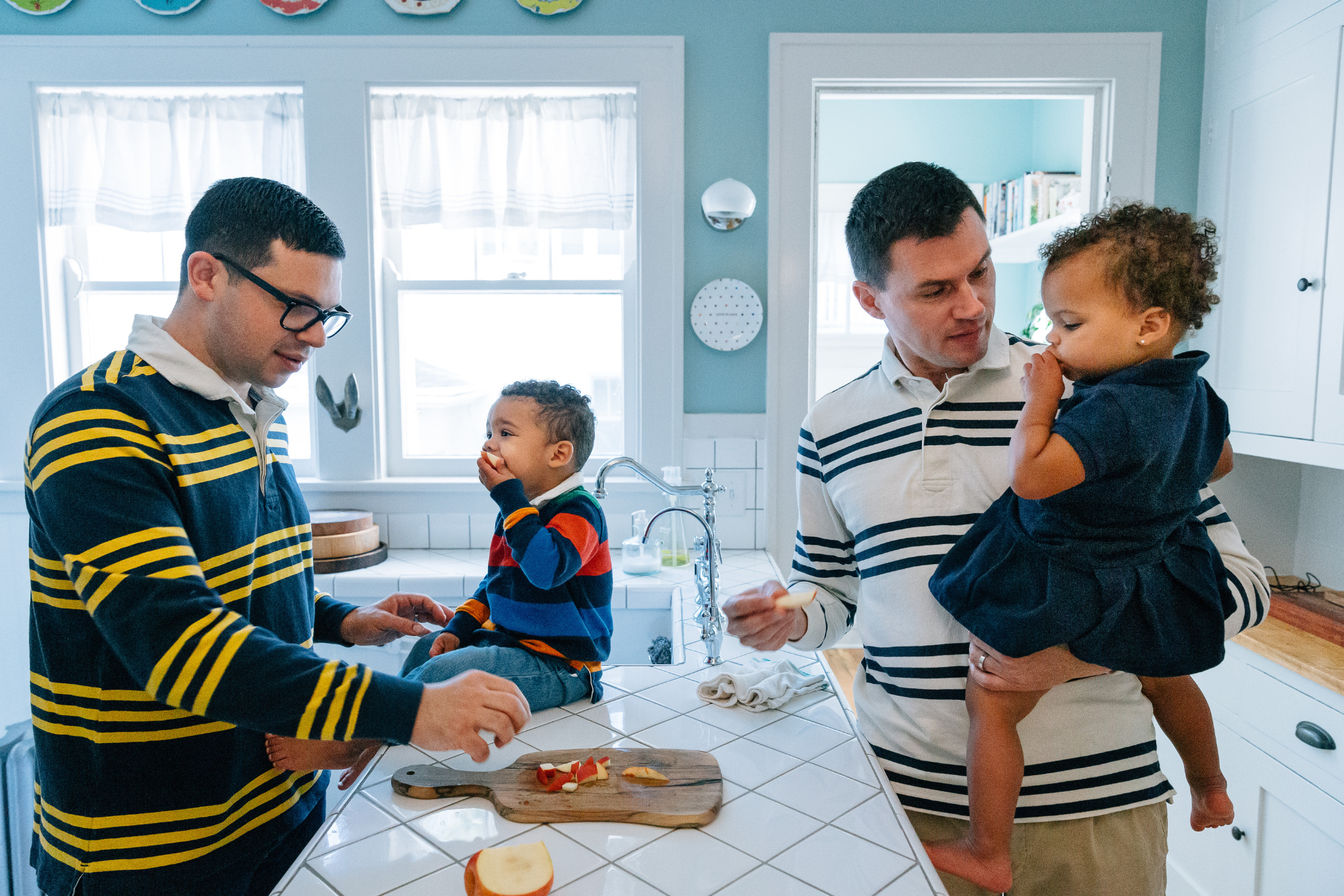 Two adult men and two young children in a bright kitchen. One man with glasses is slicing fruit on a cutting board near a boy sitting on the counter, and the other man holds a toddler.