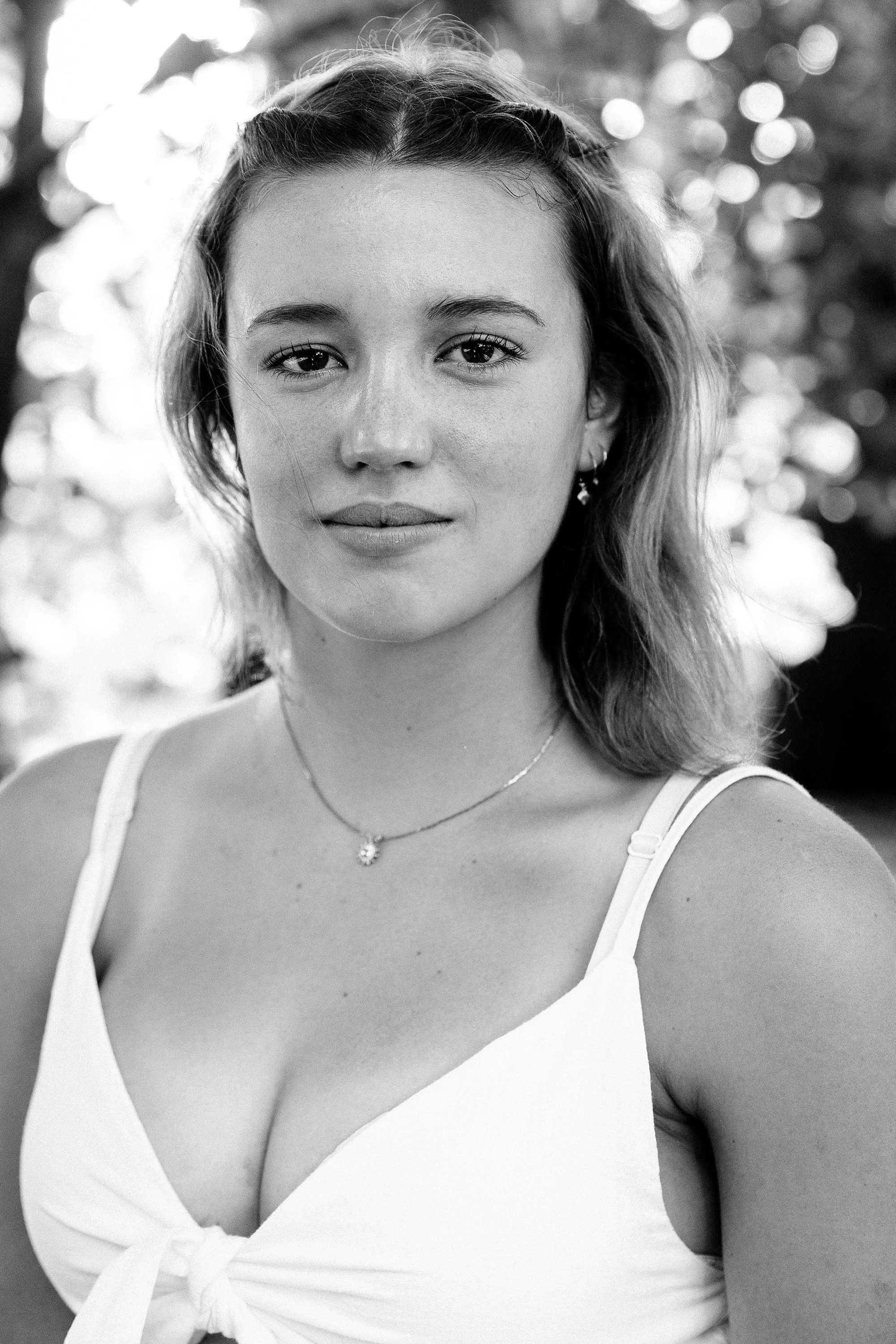 Black-and-white portrait of a young woman with light skin, shoulder-length hair, and a calm expression, wearing a tank top, necklaces, and earrings, outdoors with a blurred background of trees and sunlight.