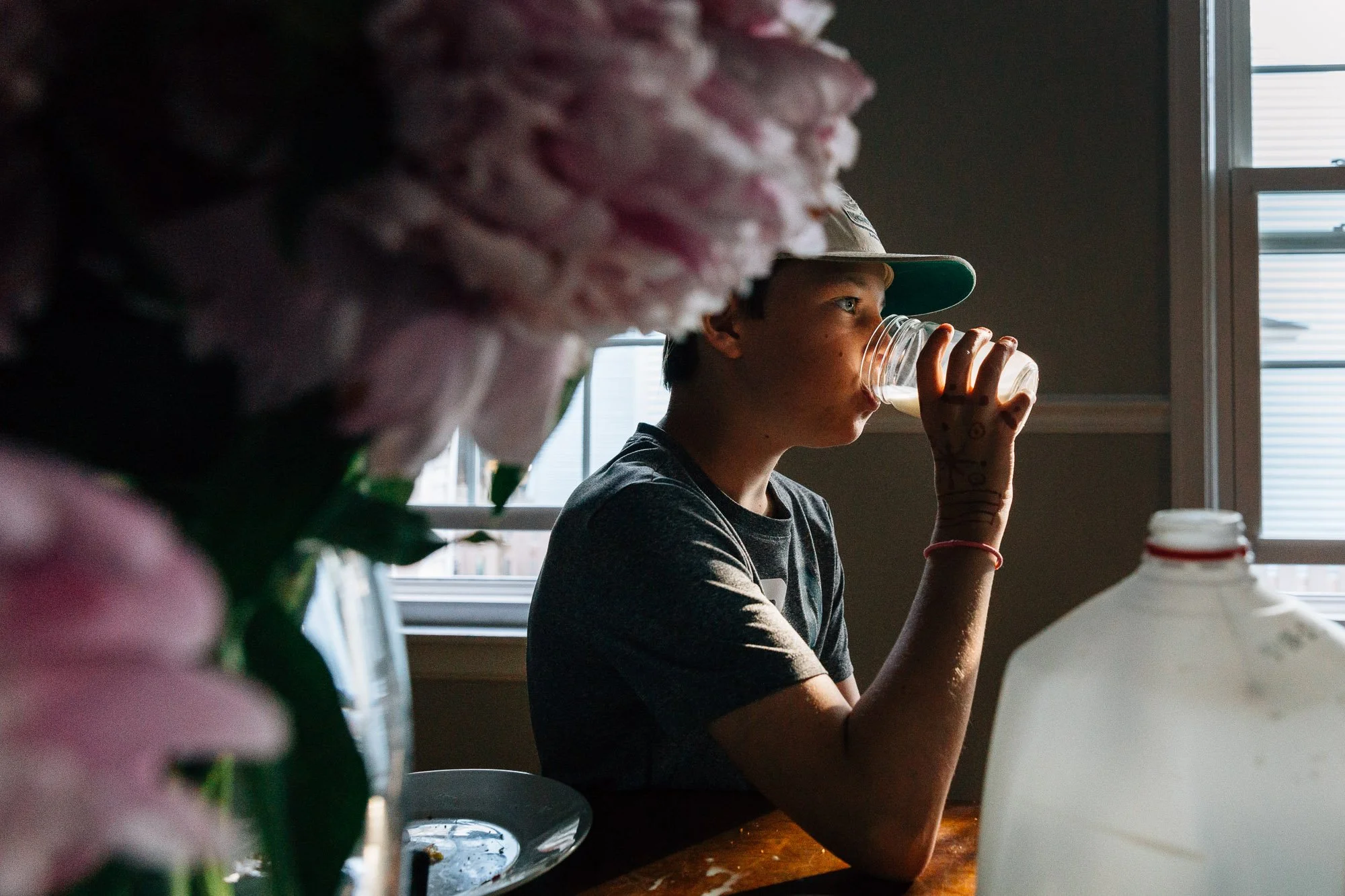 A boy with a baseball cap drinking from a glass mug at a table, with pink flowers and a milk jug in the foreground, and windows in the background.