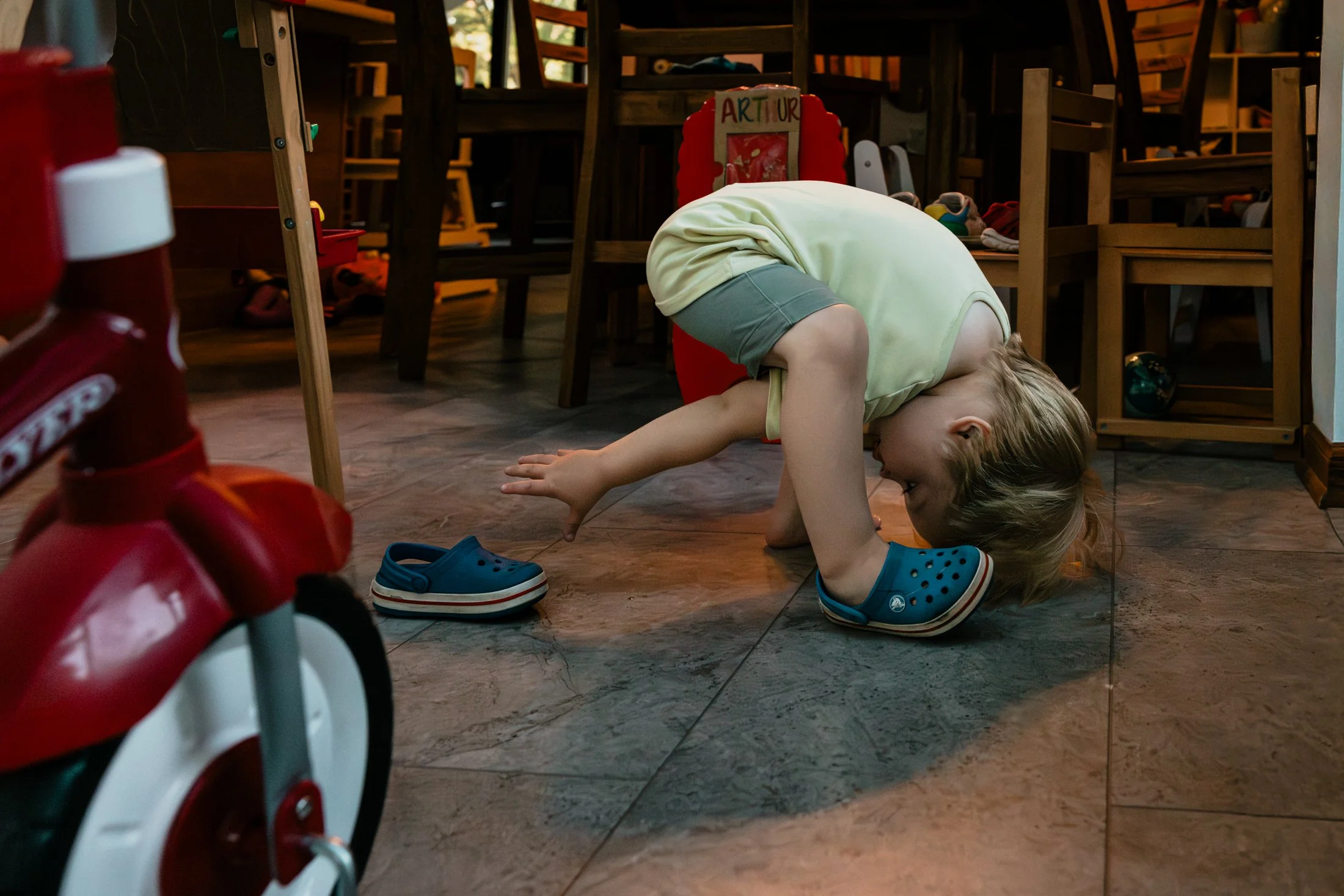 A young kneeling on the floor in a playful crawling position with her forehead and hands touching the ground, wearing a yellow shirt and green shorts, with blue Crocs nearby. She is in a room with wooden chairs, tables, and shoes scattered around, an