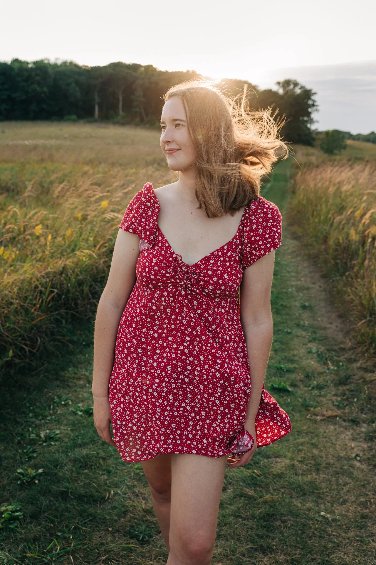 A young woman with shoulder-length red hair wearing a red dress with white floral pattern, walking along a grassy path in a field at sunset.