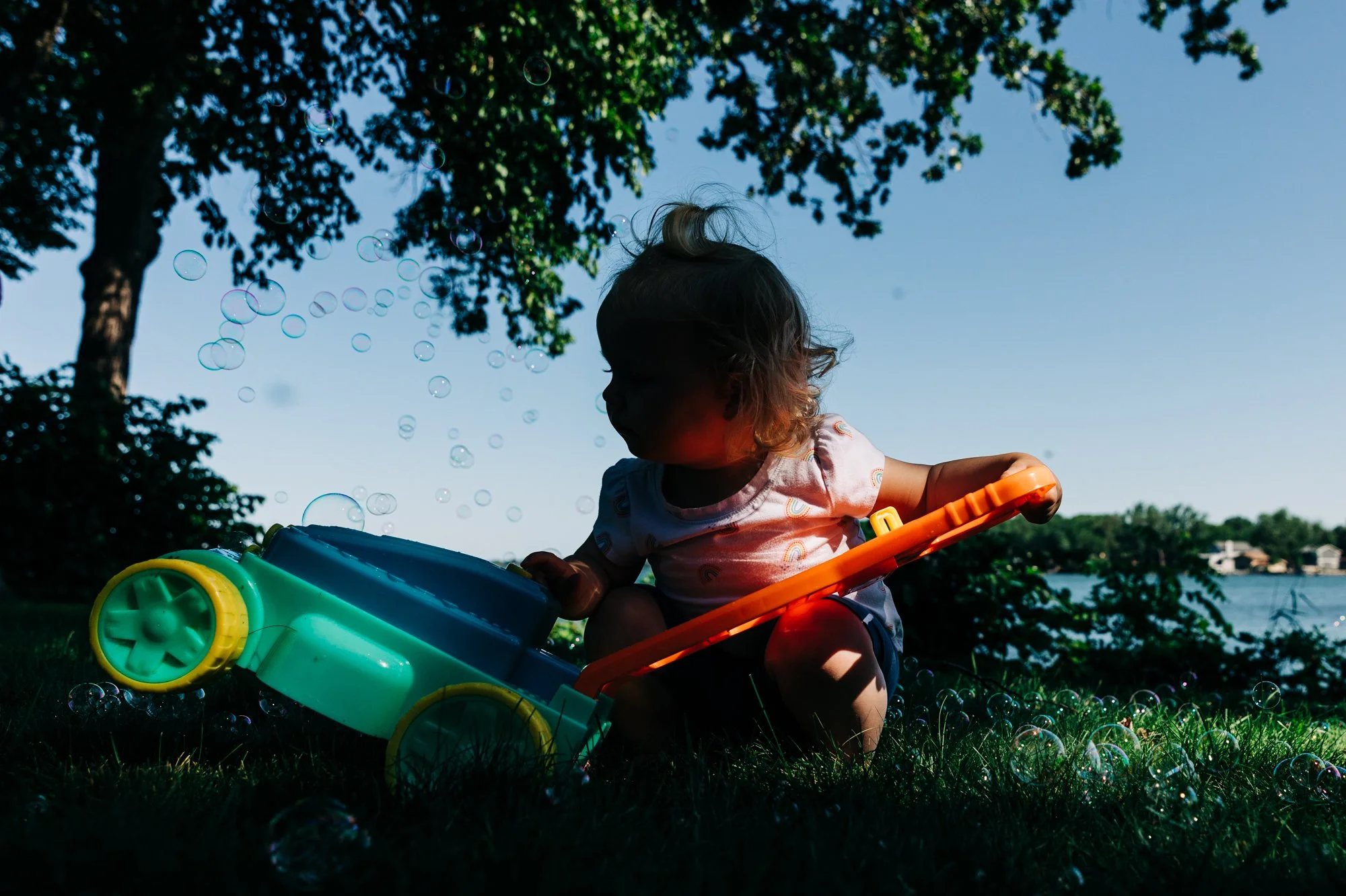 A young child sitting on the grass outdoors with bubbles floating around, near a lake or river, under a tree.