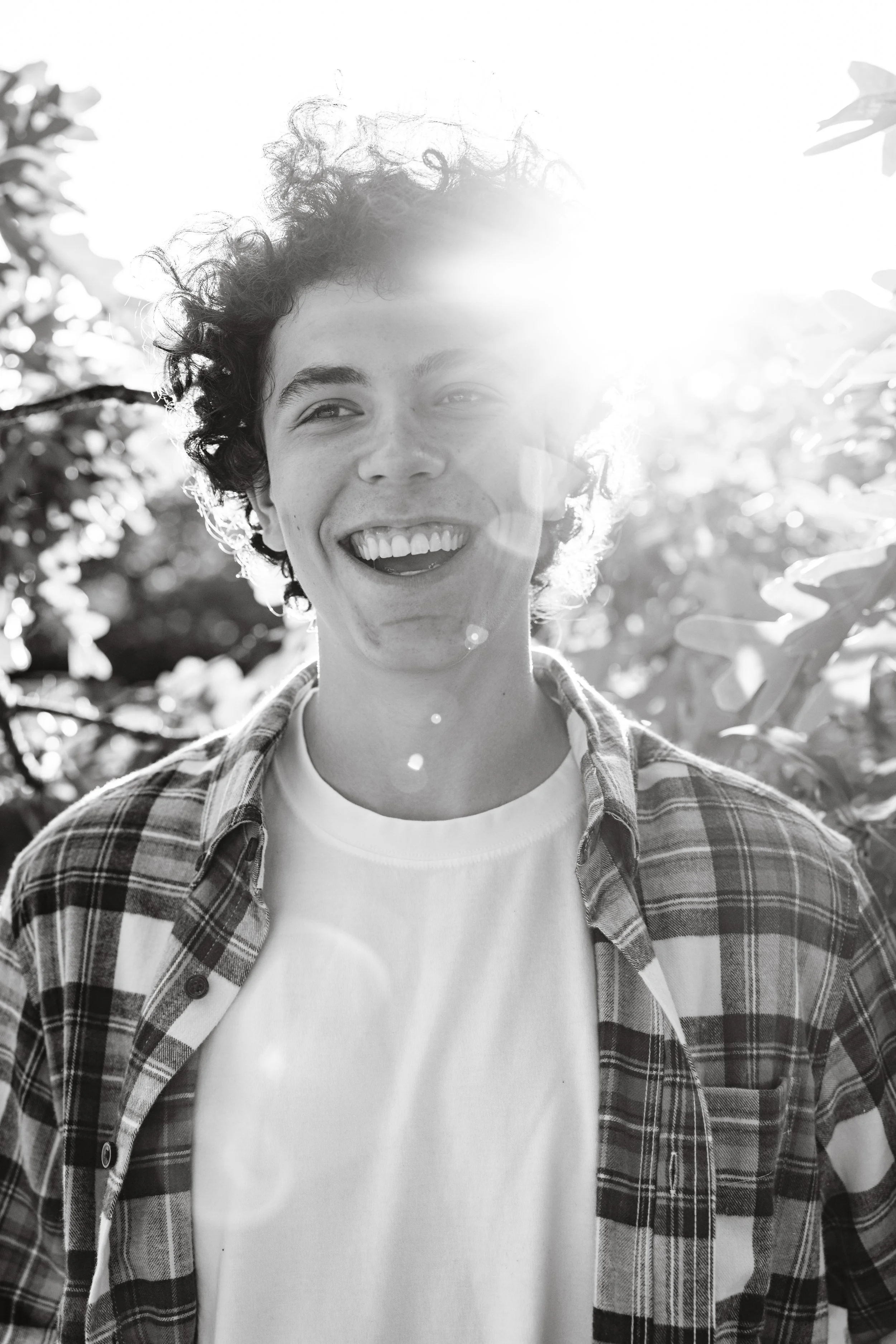 A young man with curly hair smiling outdoors, shirt unbuttoned over a white t-shirt, backlit by the sun.
