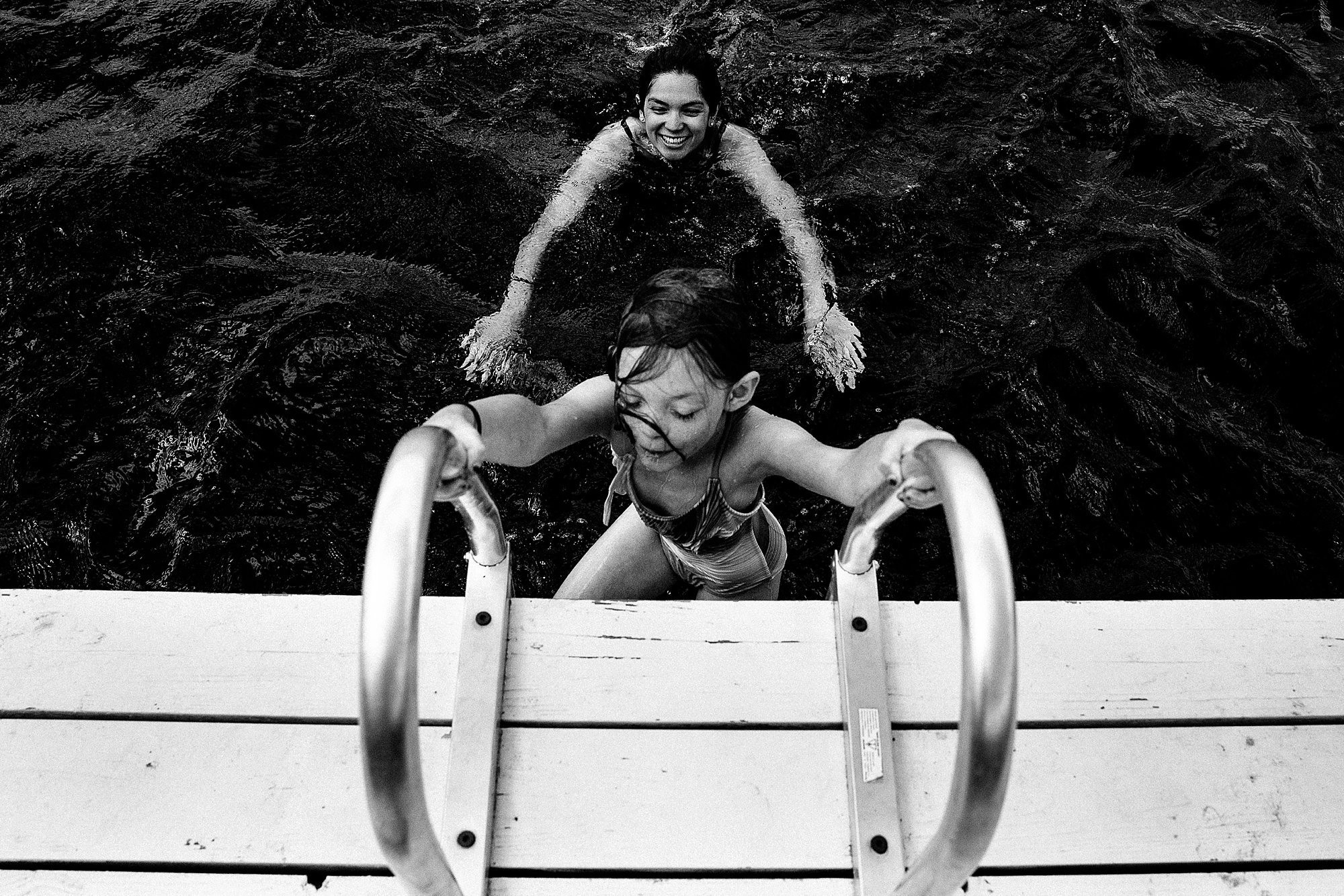 A woman and a girl swimming in a body of water, the girl is climbing out of the water using a dock ladder.