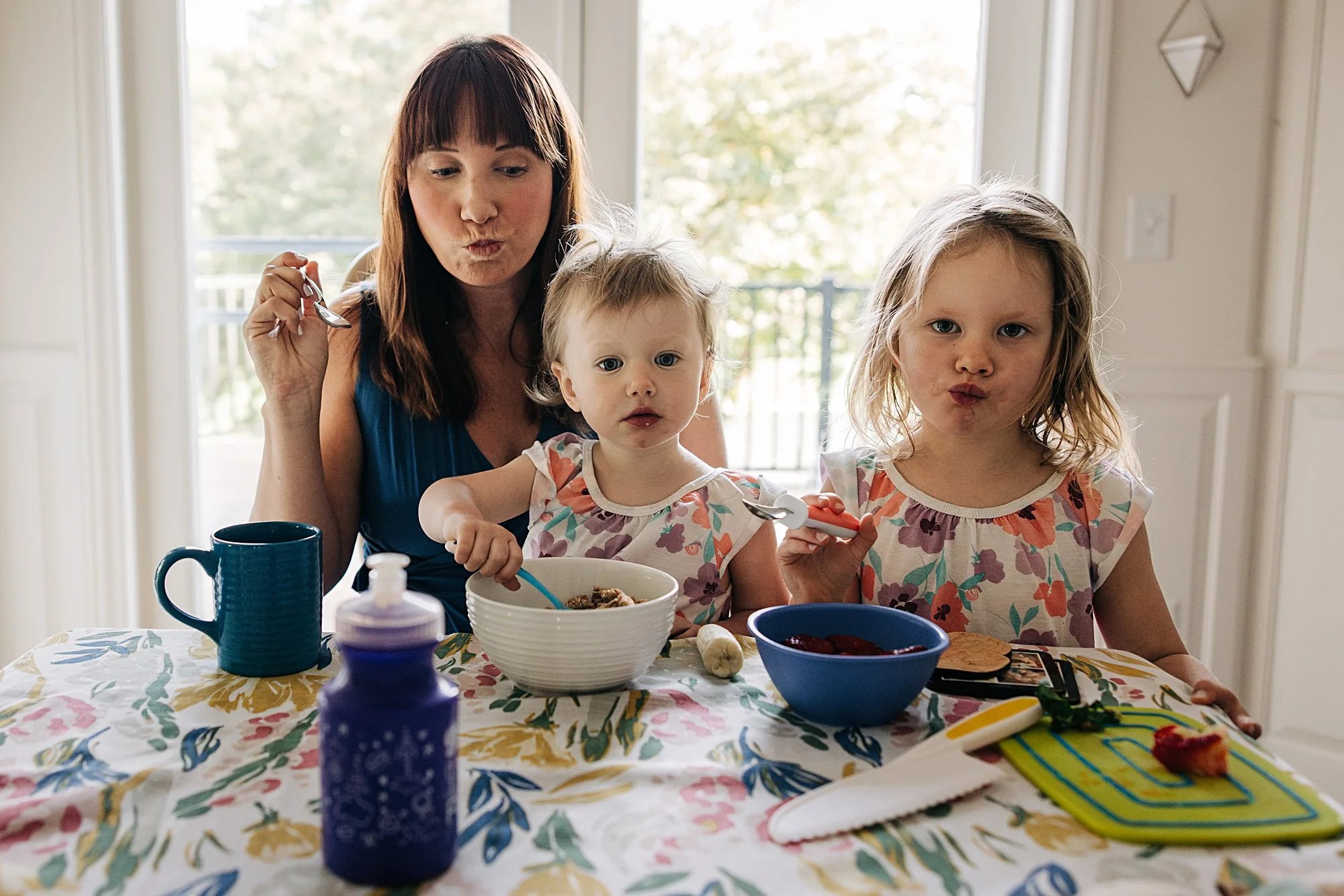 A woman and two young girls sitting at a breakfast table, making expressive faces while eating. The woman has brown hair with bangs and is wearing a blue top. The girl in the middle has light brown hair and is wearing a floral shirt, while the girl o
