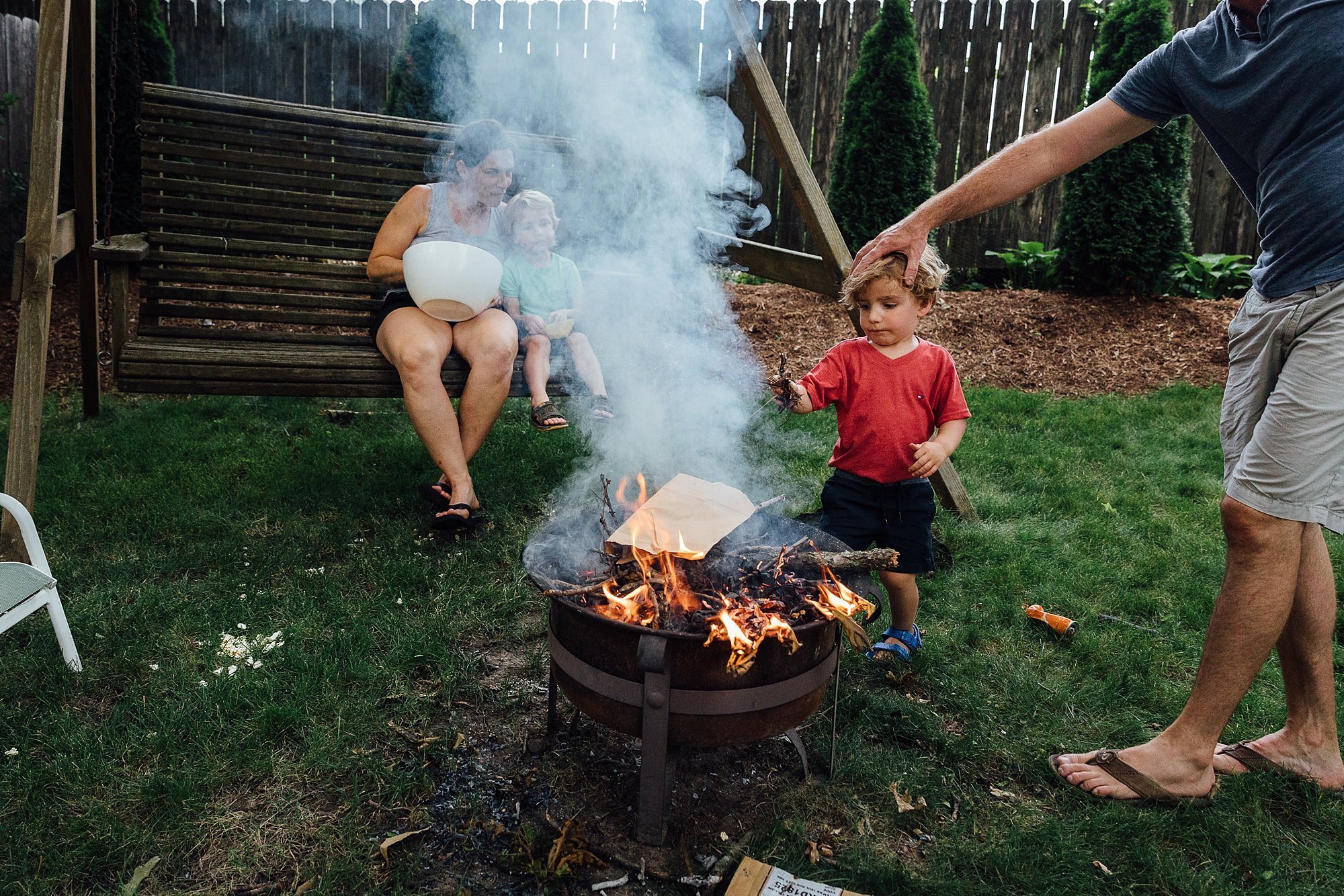 A group of people gathered around a backyard fire pit, with a woman and two children sitting on a wooden swing in the background. One child is roasting marshmallows or food over the fire, which is burning in the fire pit.