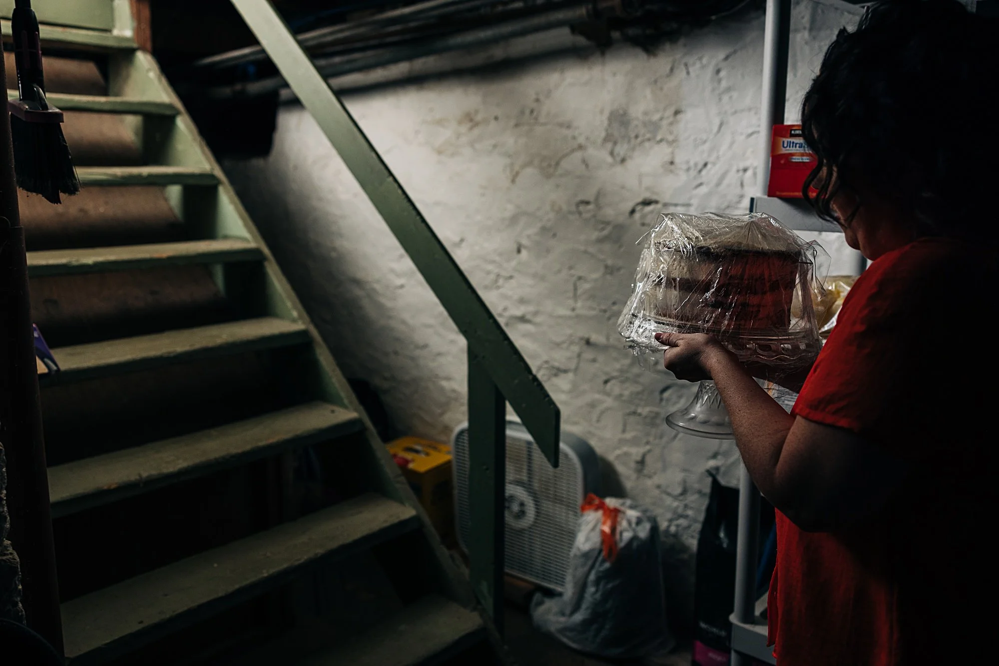 A person in a red shirt holding a cake on a glass cake stand, with a staircase, a fan, and bags in a basement or storage area.